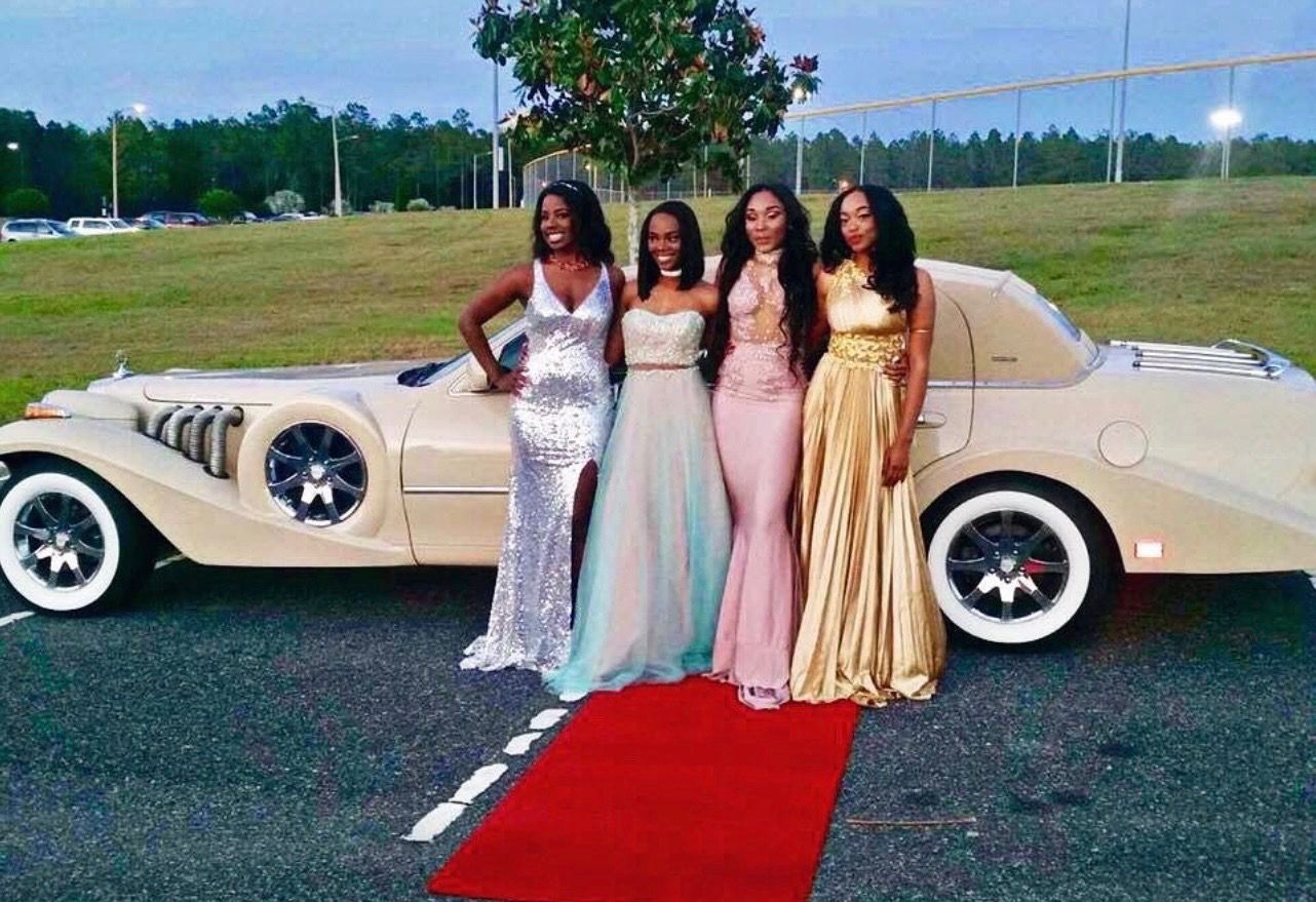 Four people in formal gowns pose together on a red carpet in front of a vintage-style beige car.