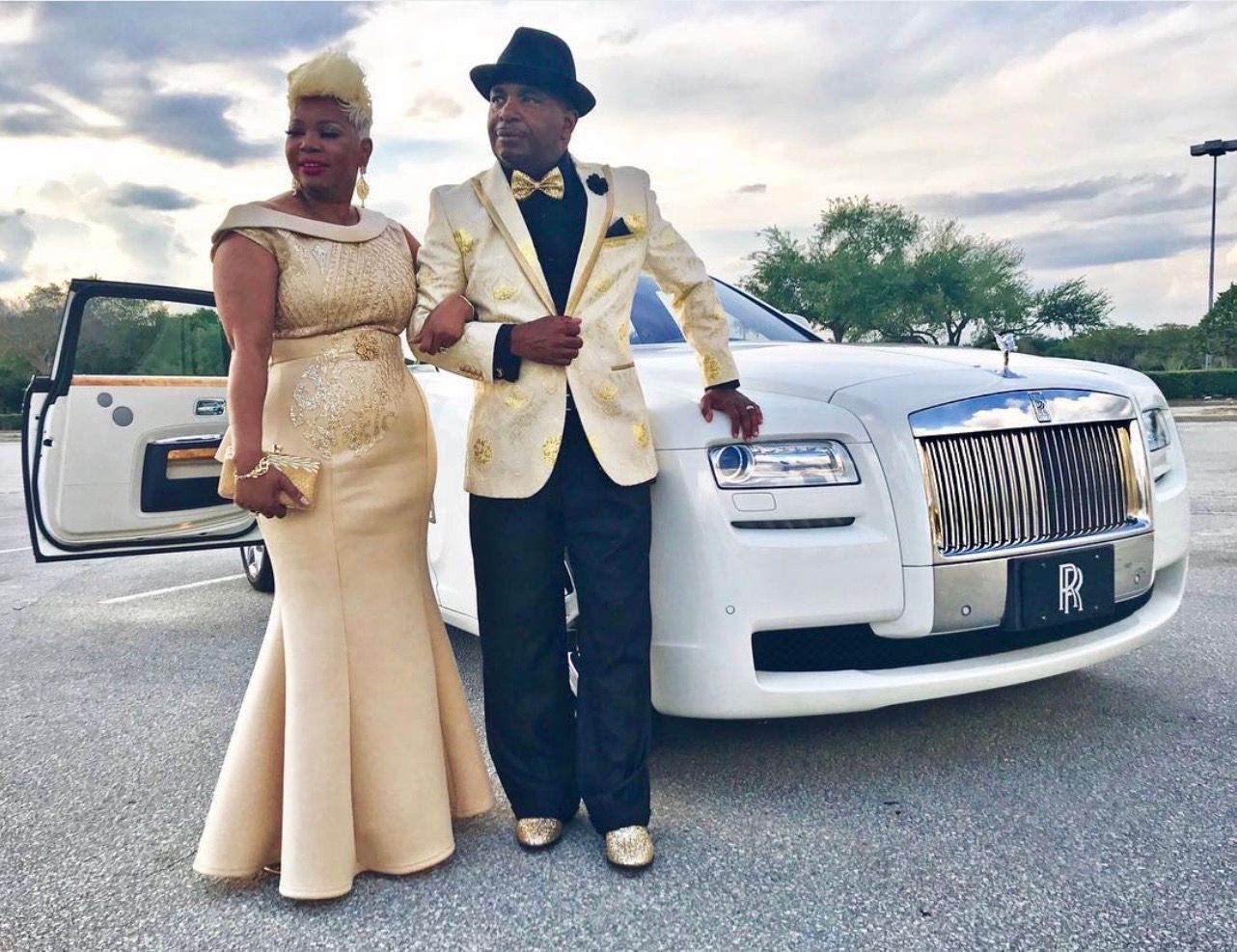 A couple in formal gold-toned evening wear stands posed together beside a white Rolls-Royce in a parking lot.