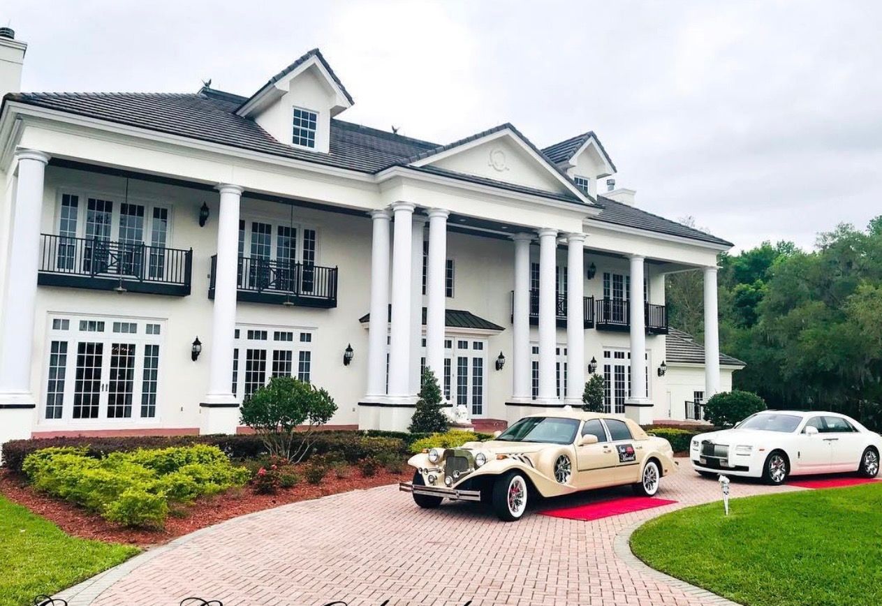 A large white mansion with columns, a brick driveway, and two luxury cars parked in front.