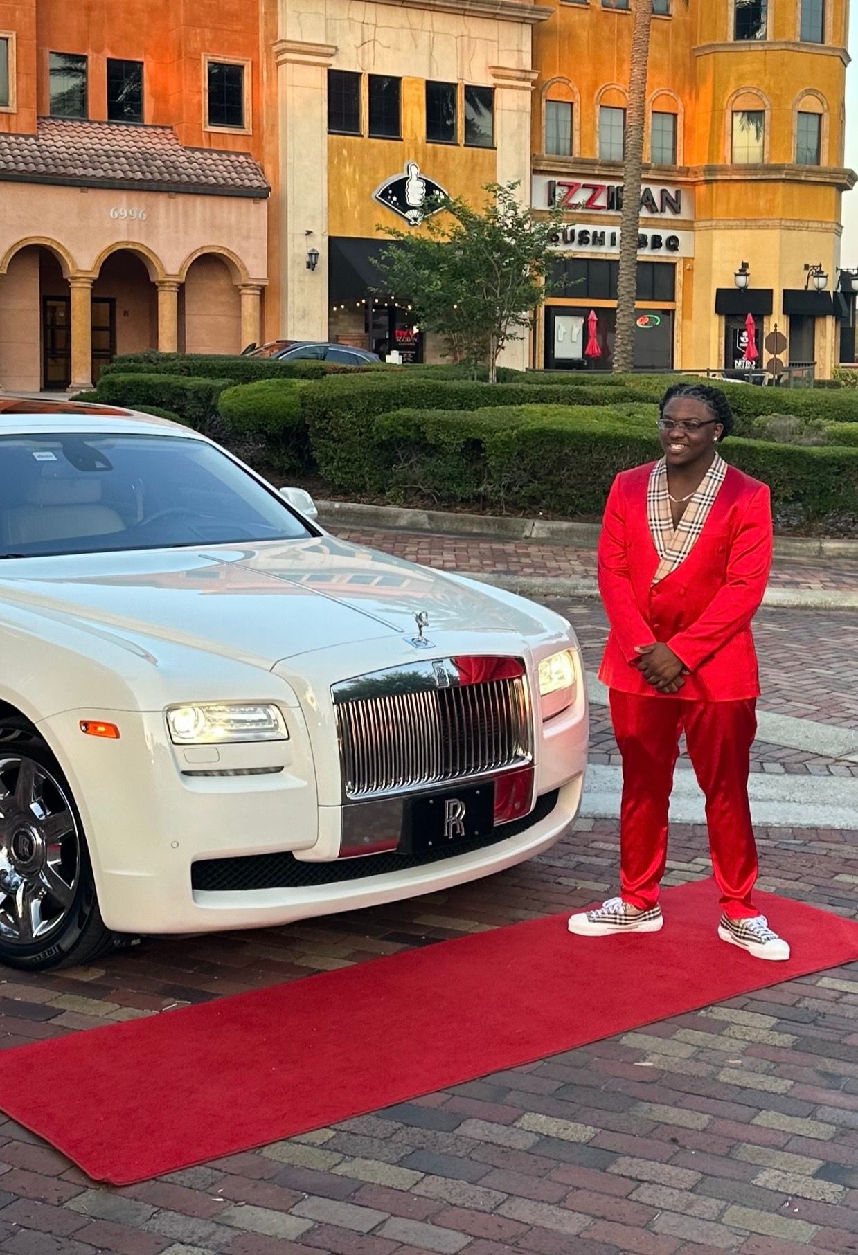 A person wearing a bright red suit stands on a red carpet next to a white Rolls-Royce in an outdoor plaza.