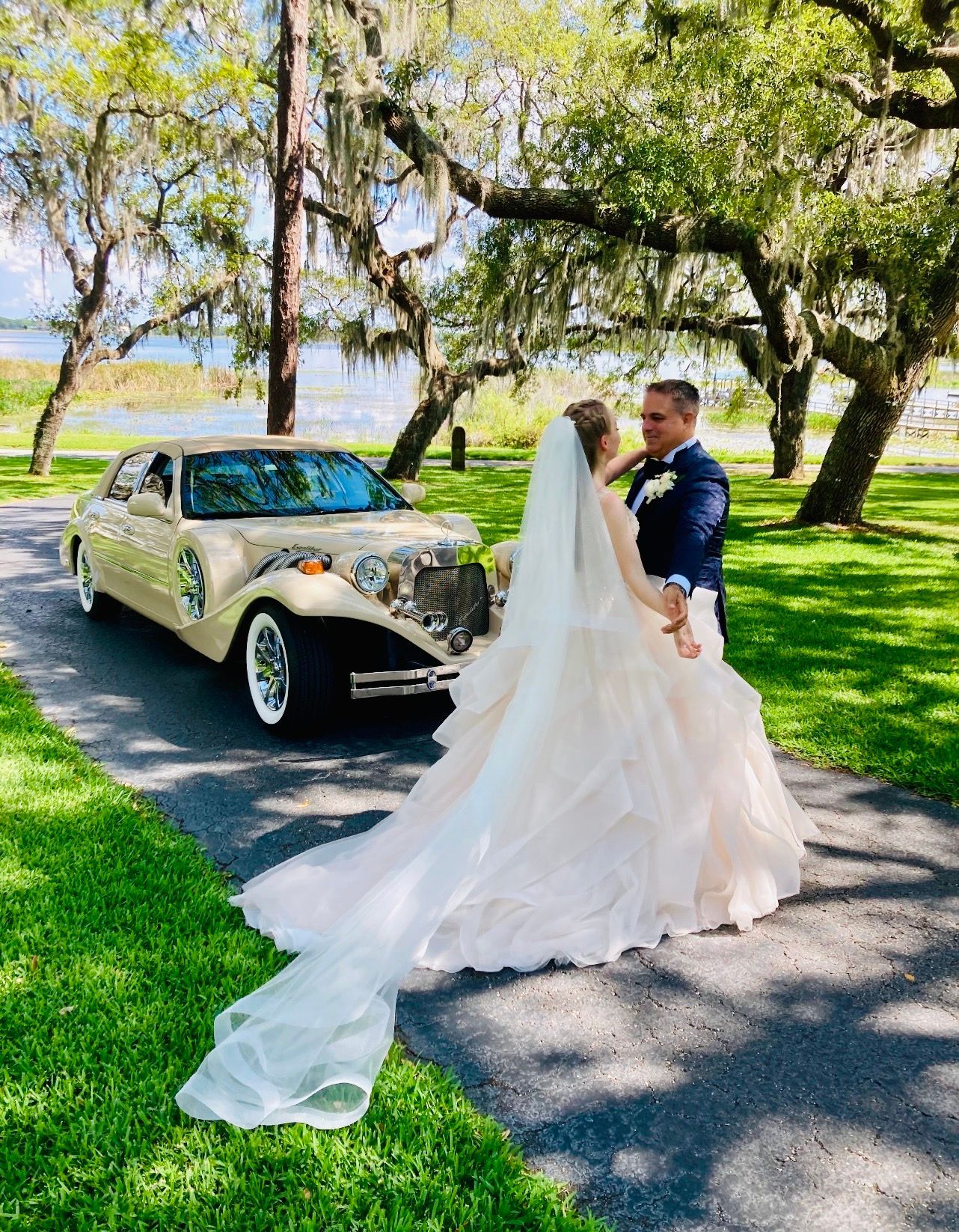 A bride and groom embrace in front of a vintage, cream-colored convertible car parked on a tree-lined driveway.