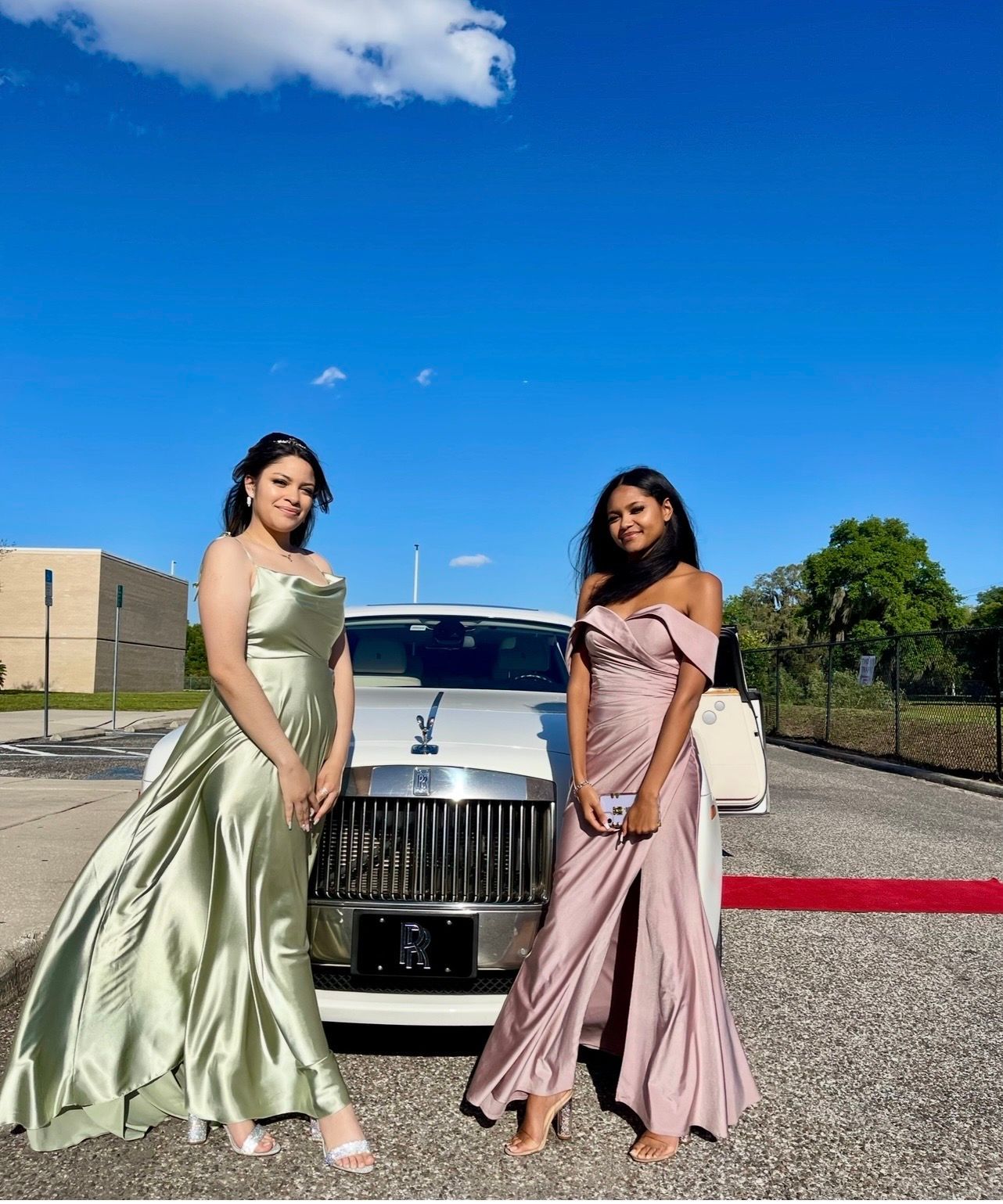 Two people in formal gowns stand in front of a white luxury car on a sunny day in a paved lot.