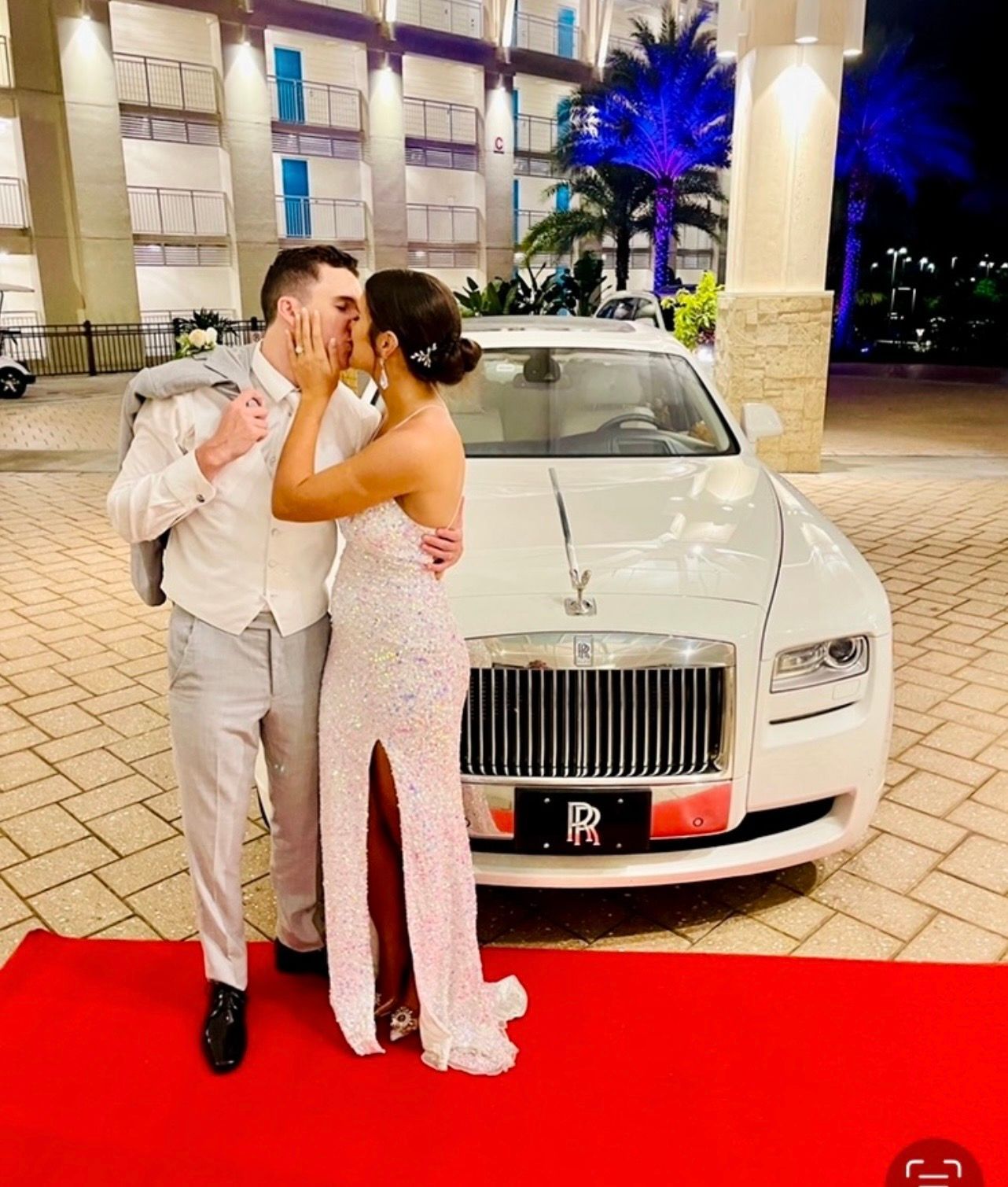 A couple kisses on a red carpet in front of a white Rolls-Royce parked at an illuminated resort entrance at night.