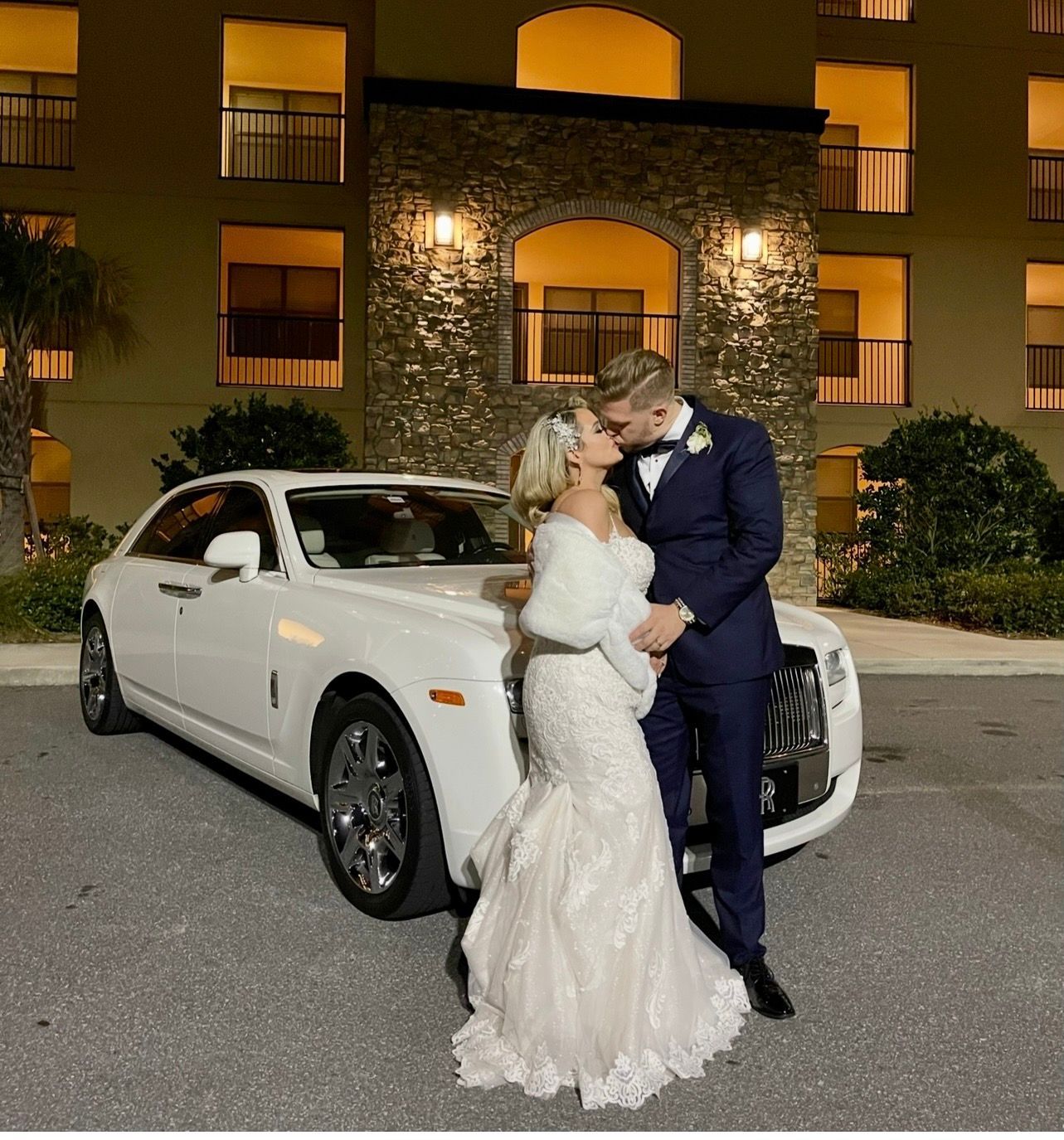 A couple in wedding attire kiss in front of a white luxury car parked outside a multi-story building at night.