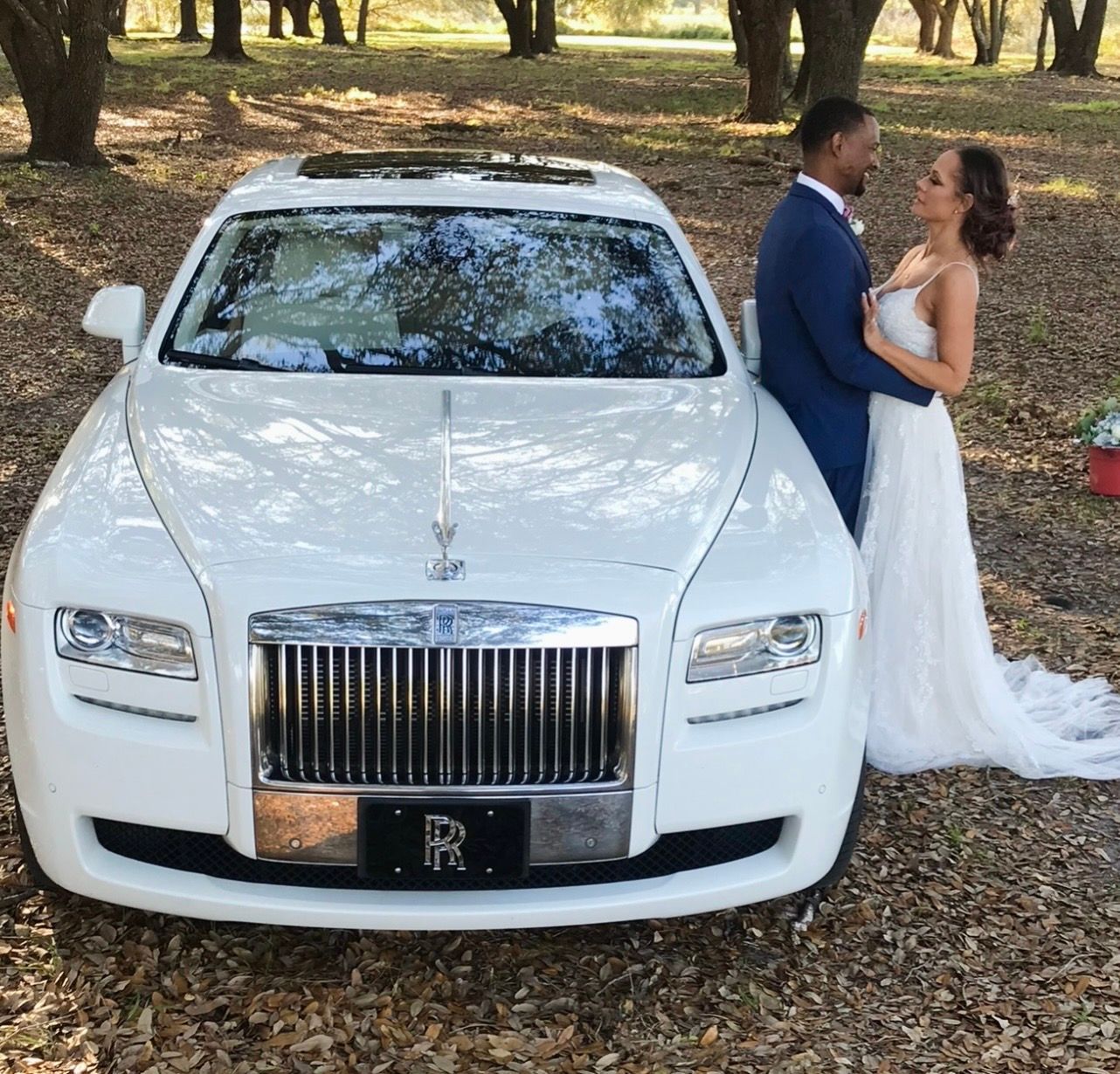 A couple in wedding attire stands beside a white Rolls-Royce parked in a wooded area covered with fallen leaves.