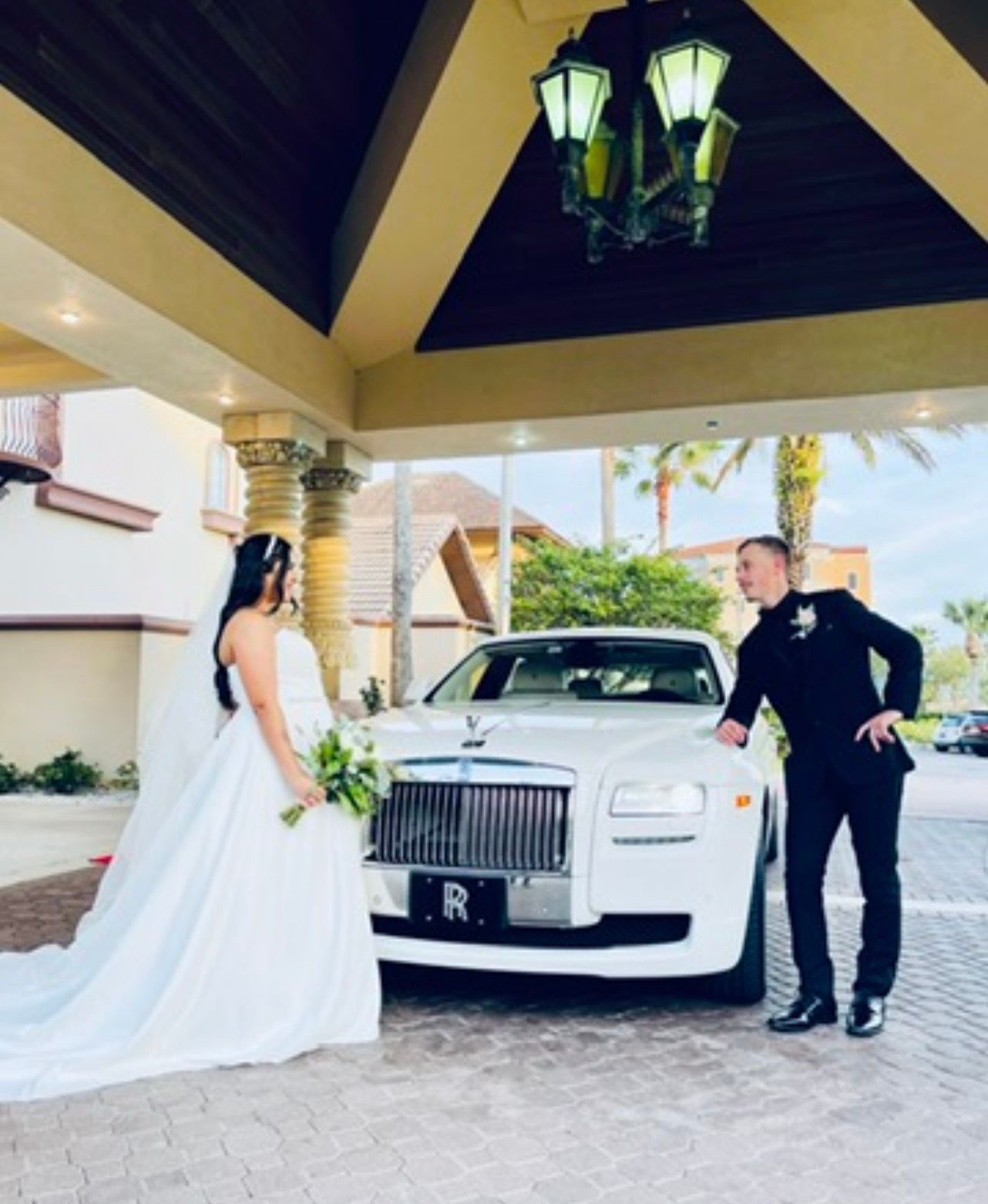 A bride and a groom in formal attire stand on either side of a white Rolls-Royce under a hotel entryway.