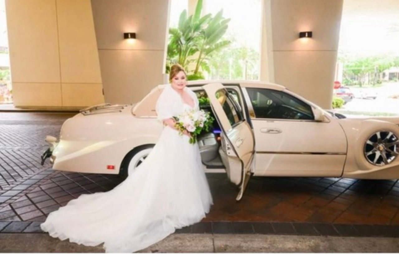 A bride holding a bouquet stands by the open door of a vintage-style, cream-colored car under a covered entrance.