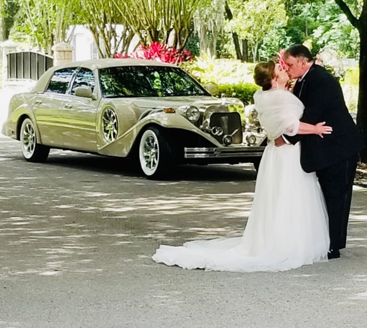 A bride and groom kiss outdoors in front of a vintage beige wedding limousine.