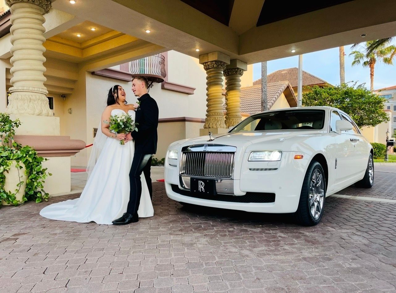 A wedding couple stands laughing next to a white Rolls-Royce parked under the entrance of a building.