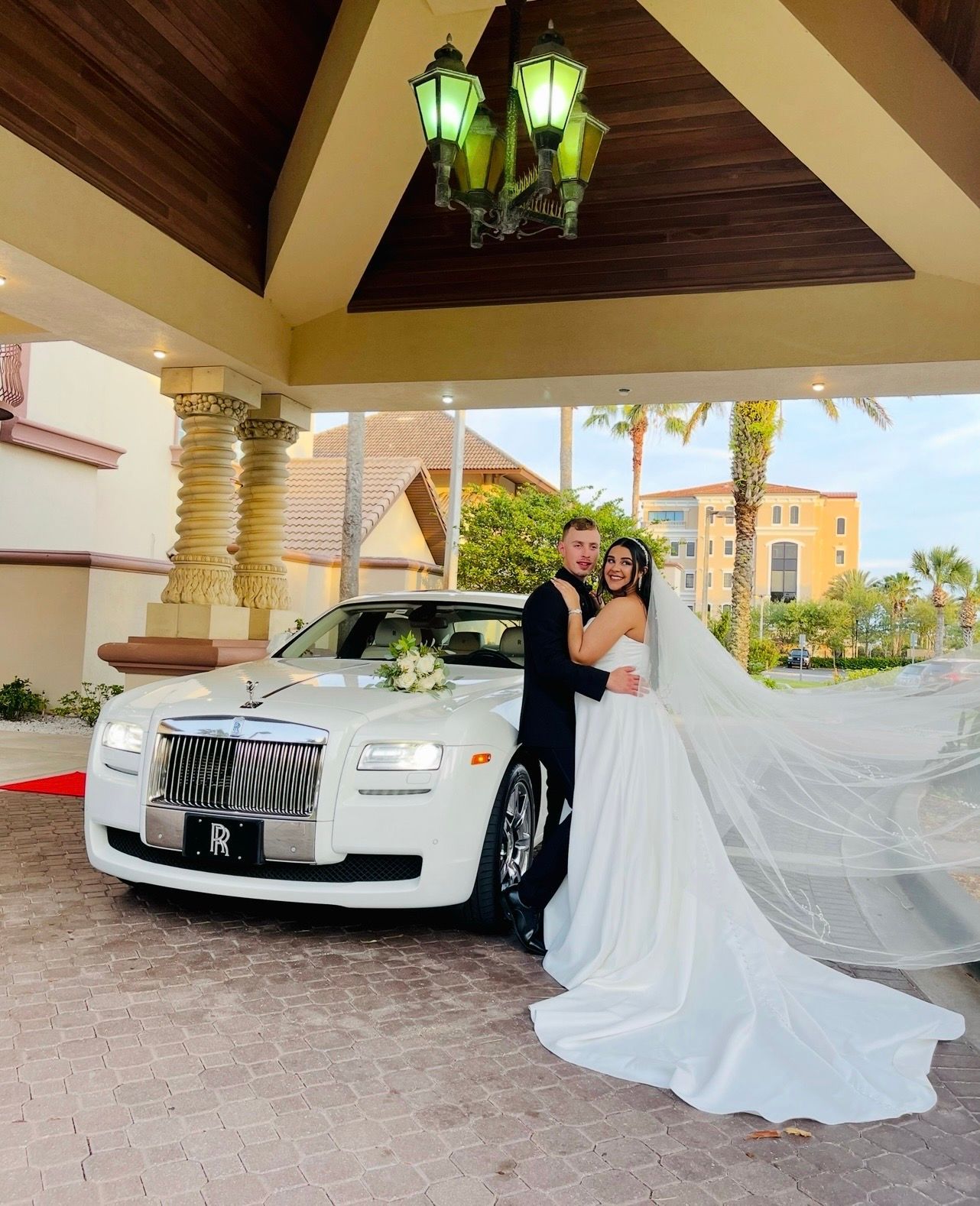 A bride and groom embrace in front of a white luxury car under a decorative covered entrance.
