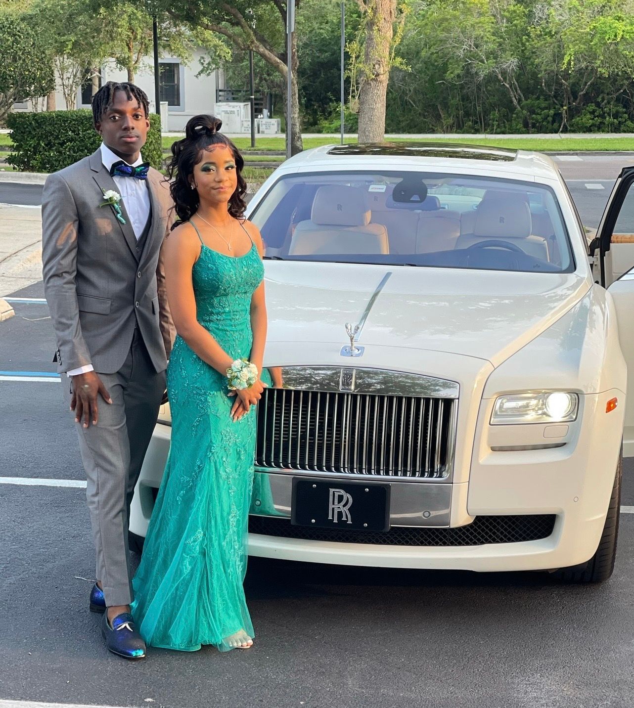 A formal couple poses in front of a white Rolls-Royce in a parking lot.