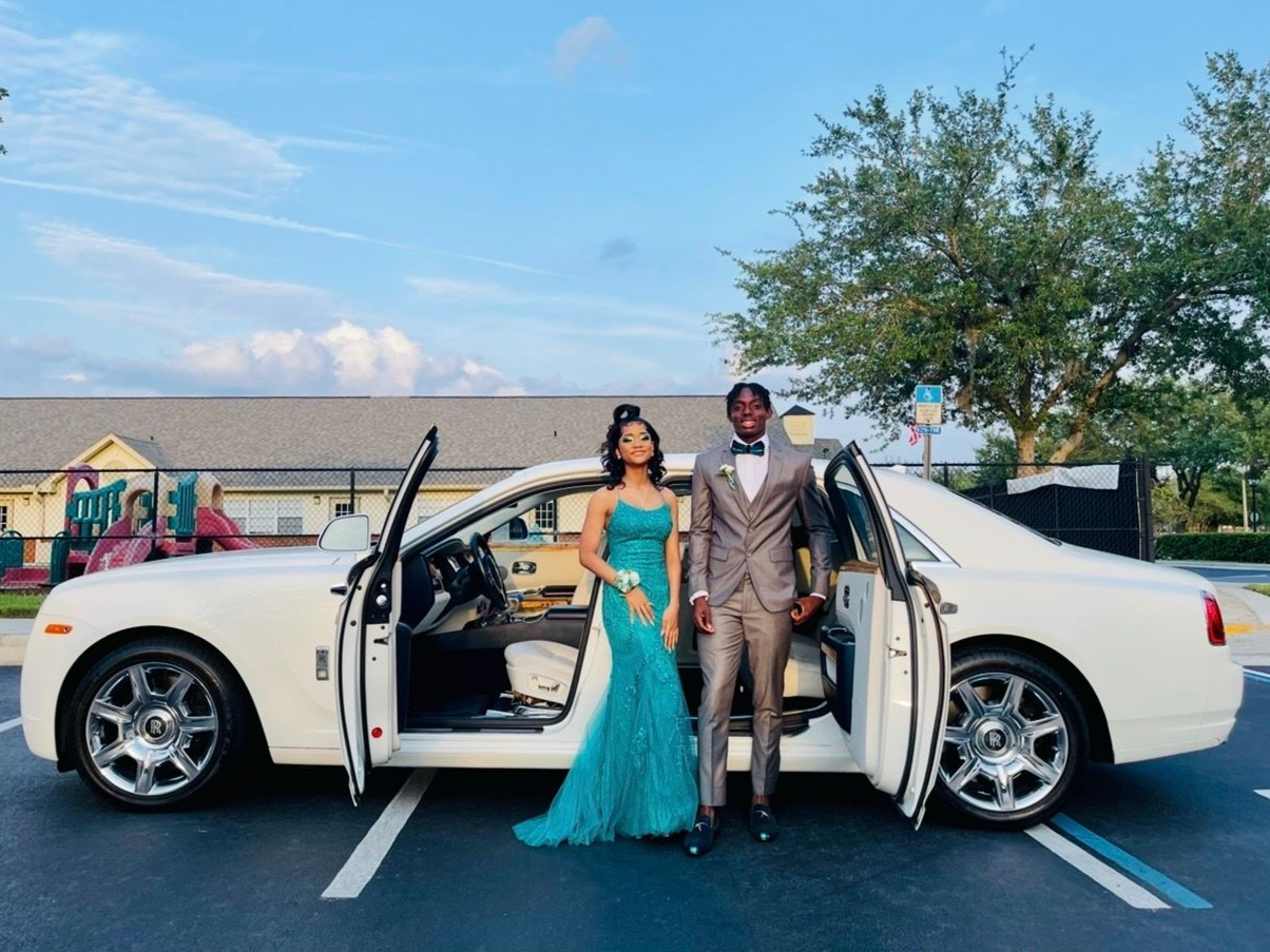 A couple in formal attire stands by an open white luxury car in a parking lot during sunset.