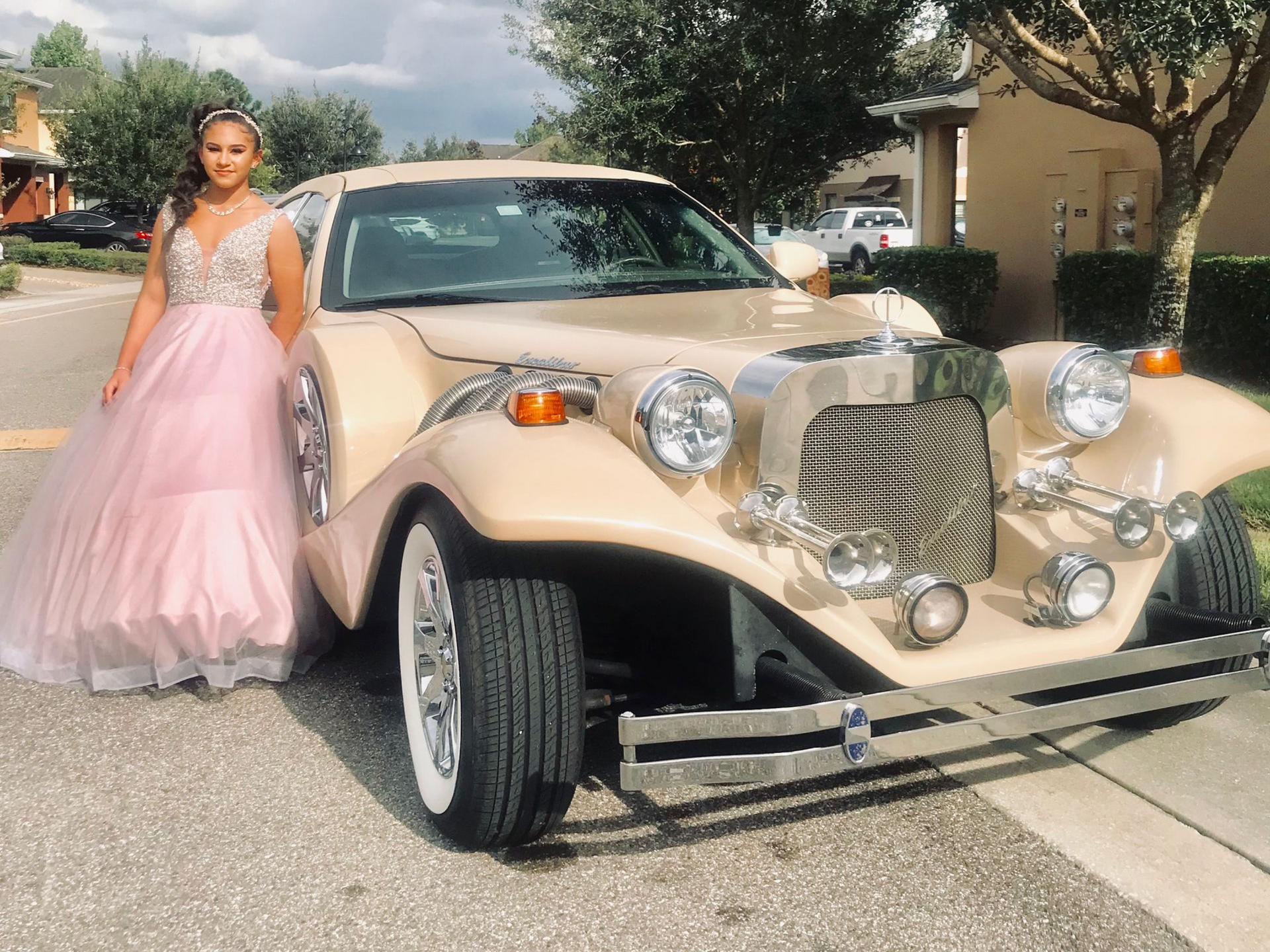 A person in a long pink gown poses next to a vintage-style tan car parked on an asphalt street.