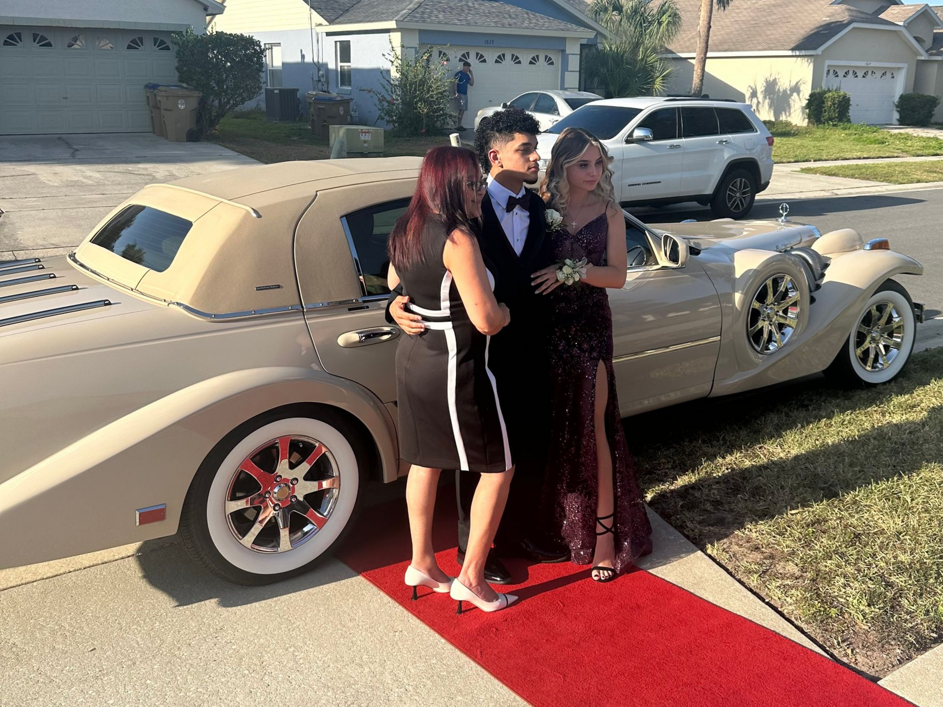 A student in a tuxedo and a companion in a sequined gown stand on a red carpet next to a vintage-style car in a driveway.