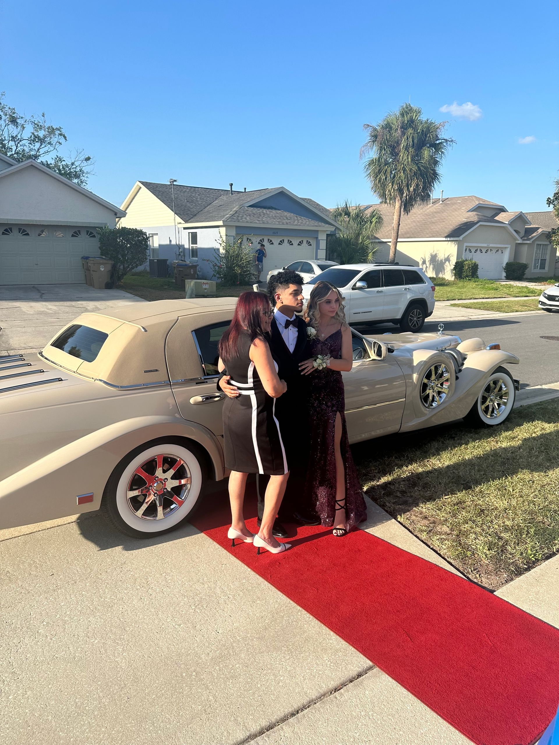 Three people stand on a red carpet next to a vintage cream-colored car in a residential driveway on a sunny day.