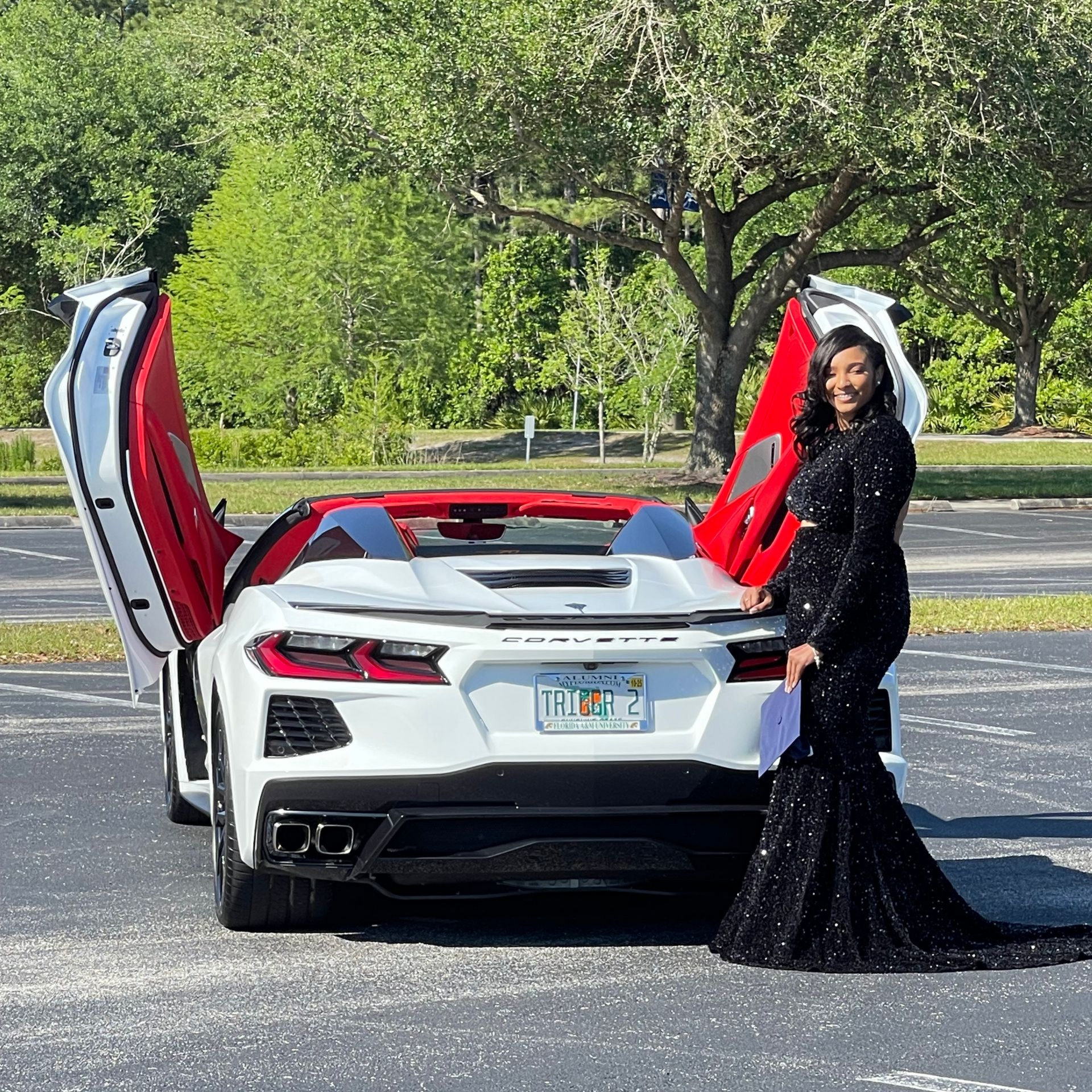 A person in a floor-length black sequined gown stands beside a white sports car with open doors in a sunny parking lot.