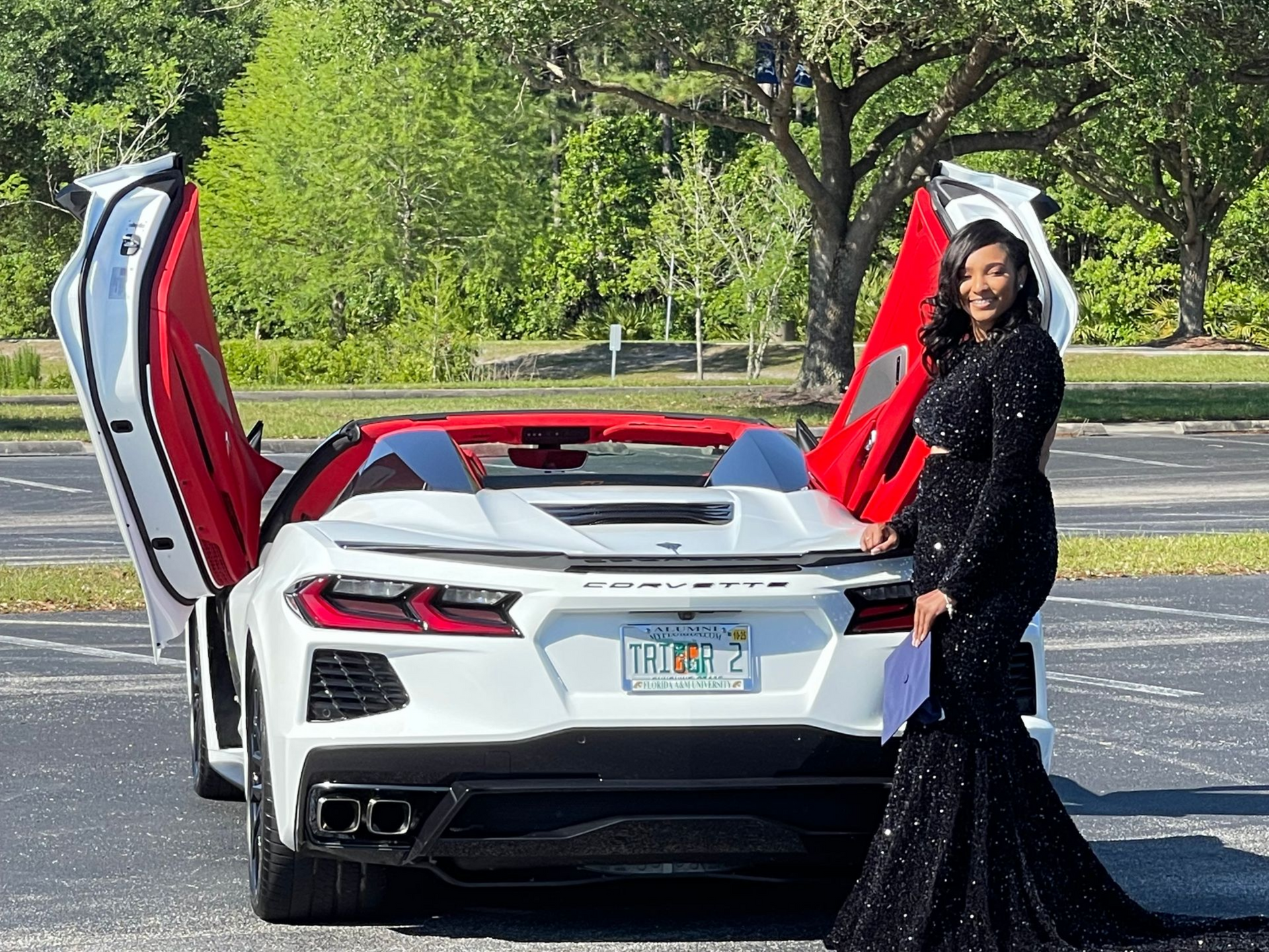 A smiling person in a sparkling black gown stands beside a white Corvette with red interior and open butterfly doors.