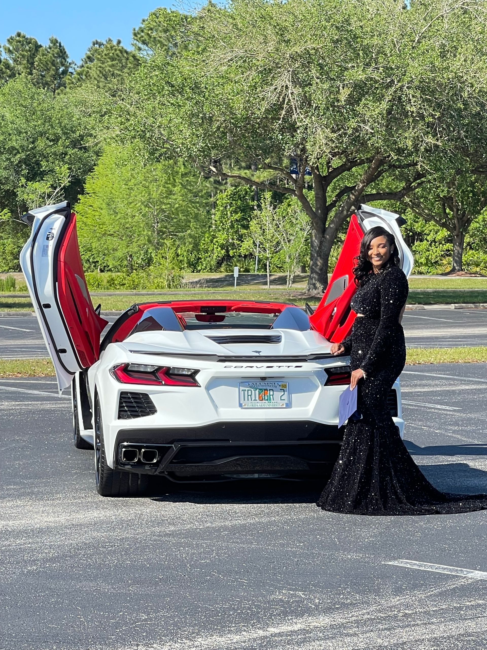 A person in a long, black sequined gown stands beside a white sports car with open doors in a paved parking lot.