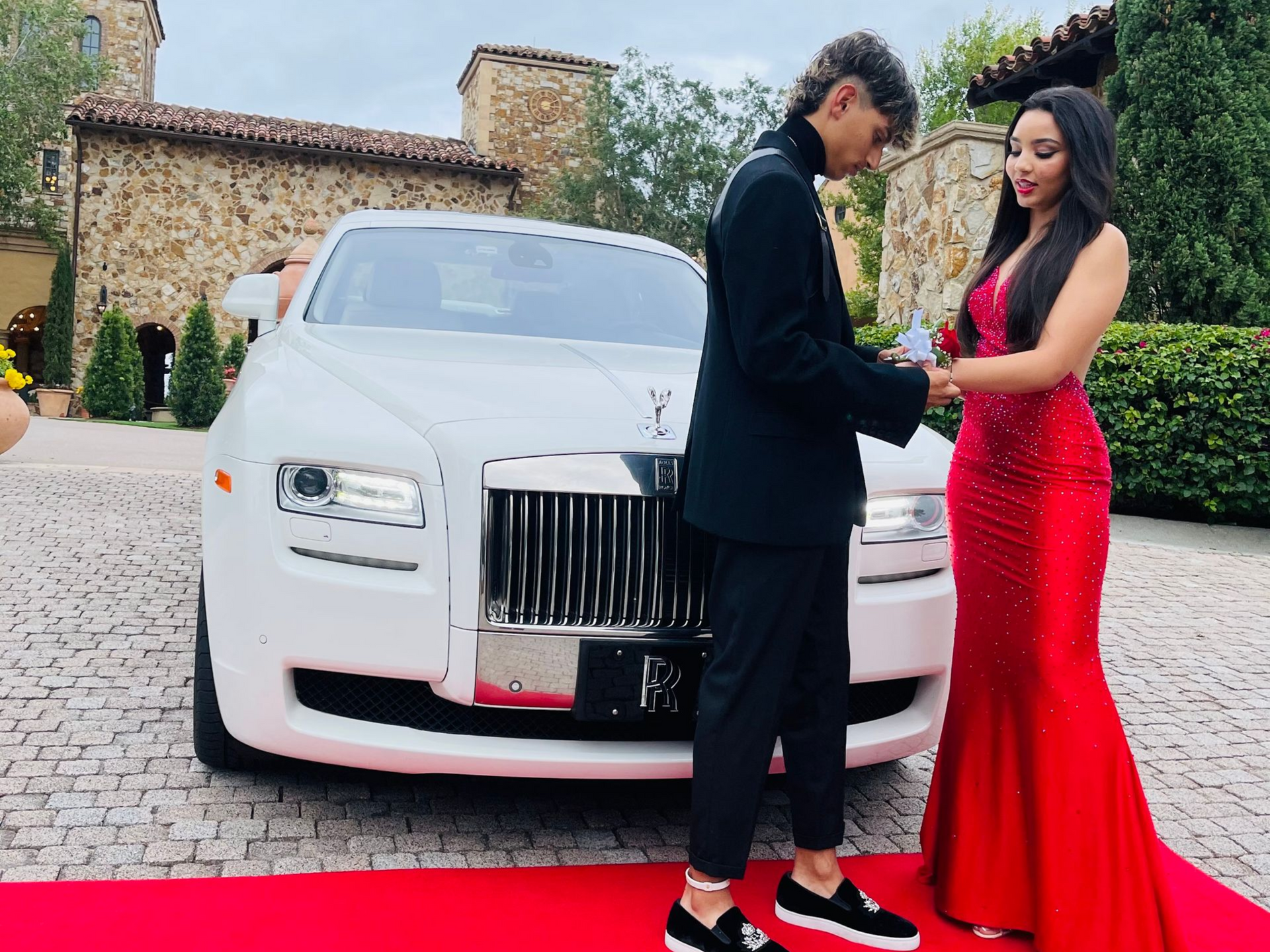 A person in a black suit places a corsage on a person in a red gown on a red carpet in front of a white Rolls-Royce.