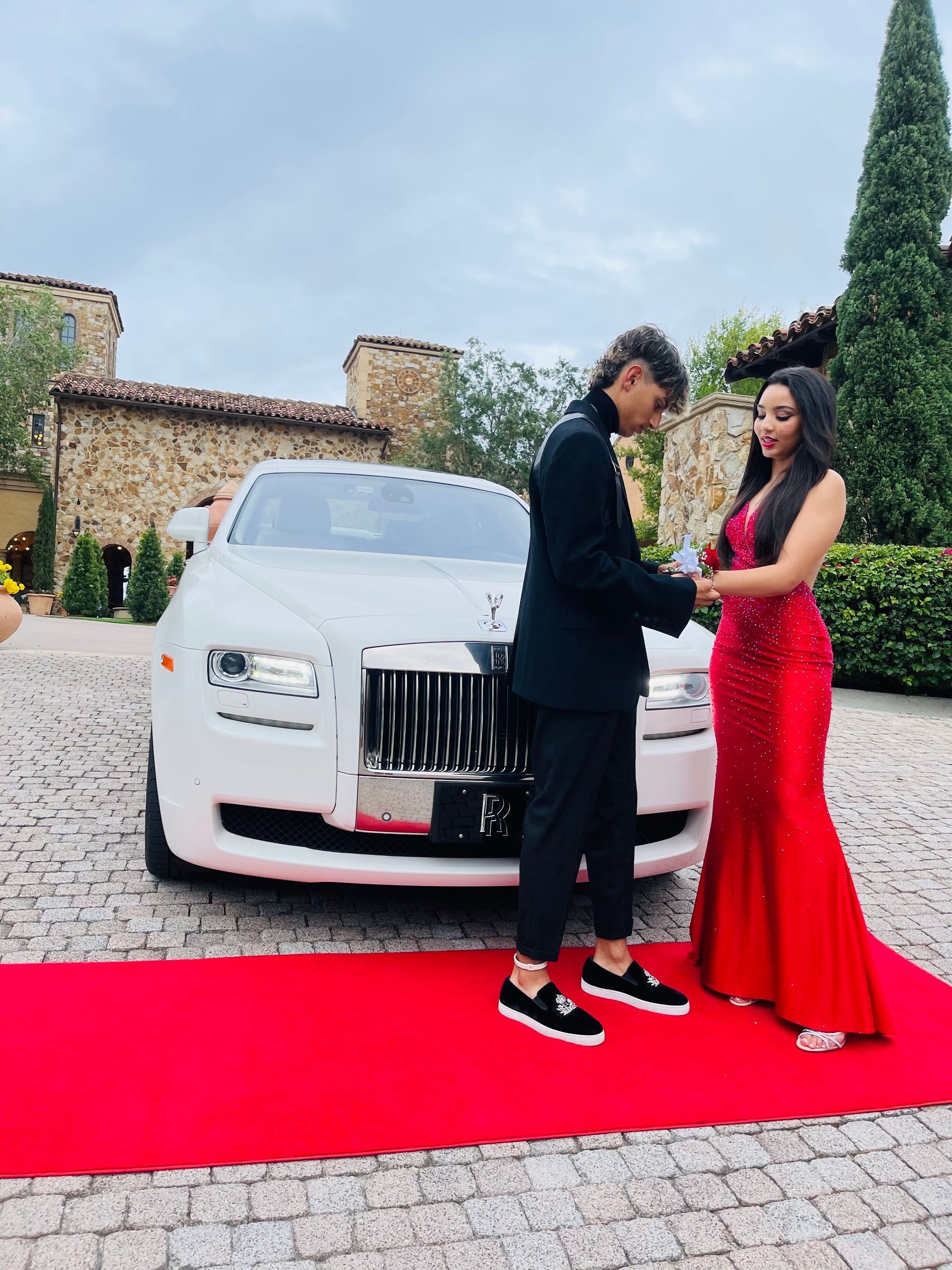 A person in a black suit places a corsage on the wrist of someone in a red gown on a red carpet in front of a white car.