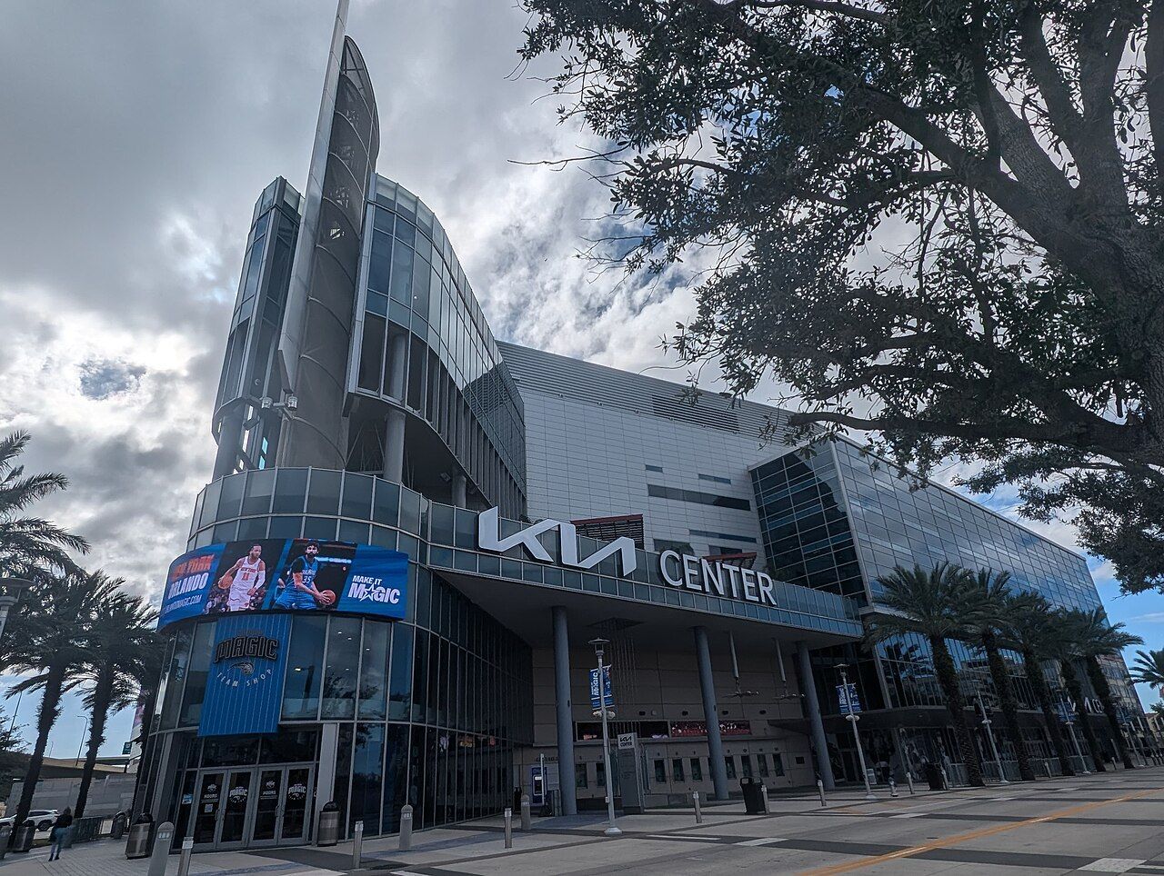 The Kia Center in Orlando, Florida, featuring a modern glass and steel exterior with palm trees on a partly cloudy day.