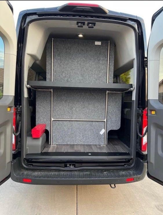 The open rear doors of a van reveal a gray, custom-built storage unit with a flat platform shelf installed in the cargo bay.