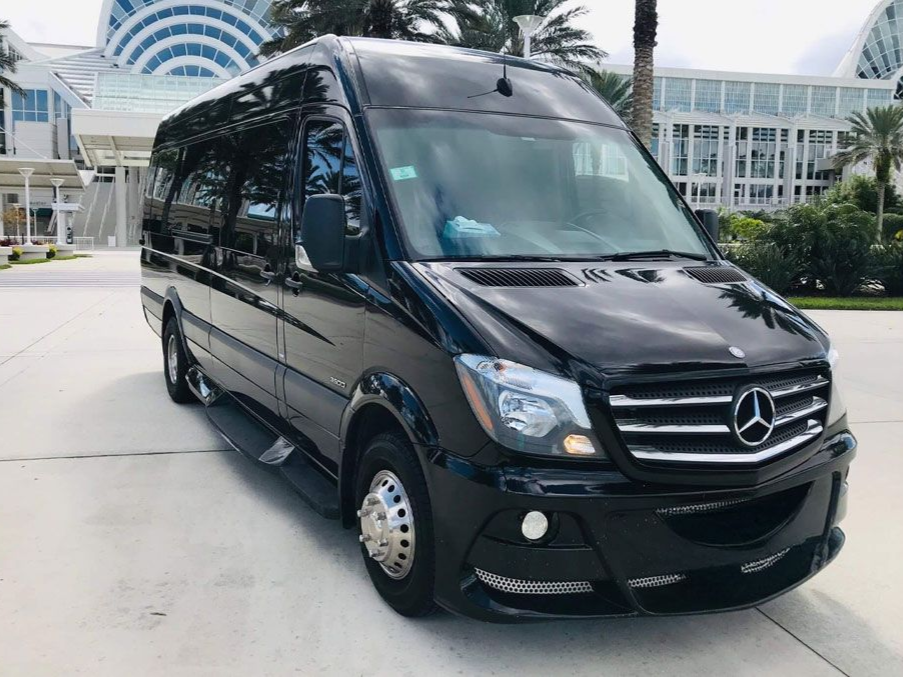 A black Mercedes-Benz Sprinter passenger van parked in front of a large, modern building with palm trees.