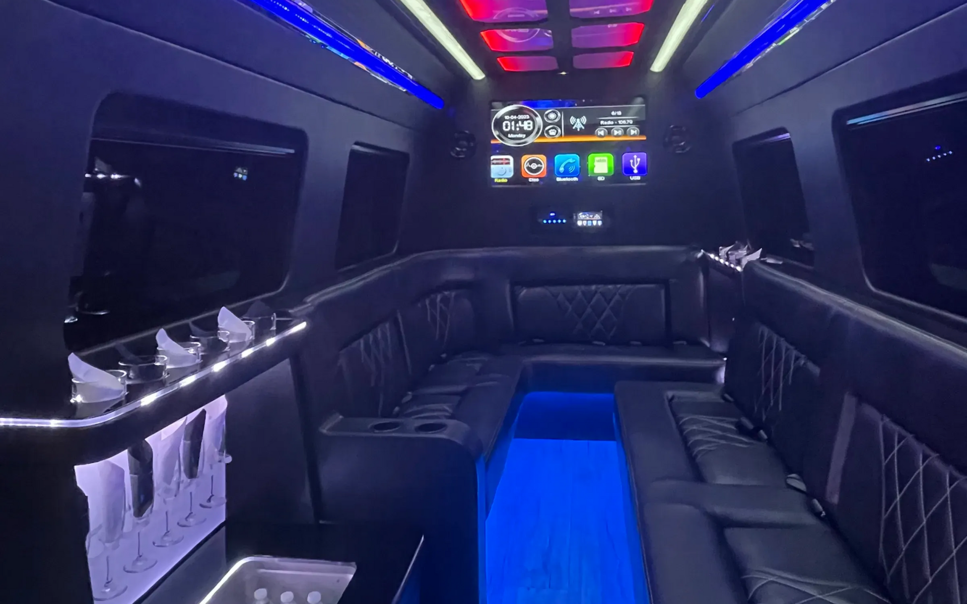 Interior of a luxury black van with blue and white lights, leather seating, a TV, and a bar area.