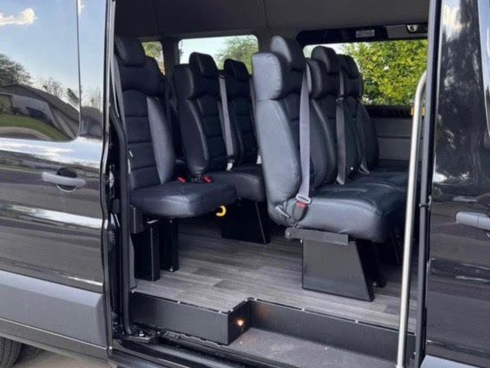 Interior of a black van featuring rows of black leather seats, a dark wood-look floor, and an open side door with a step.