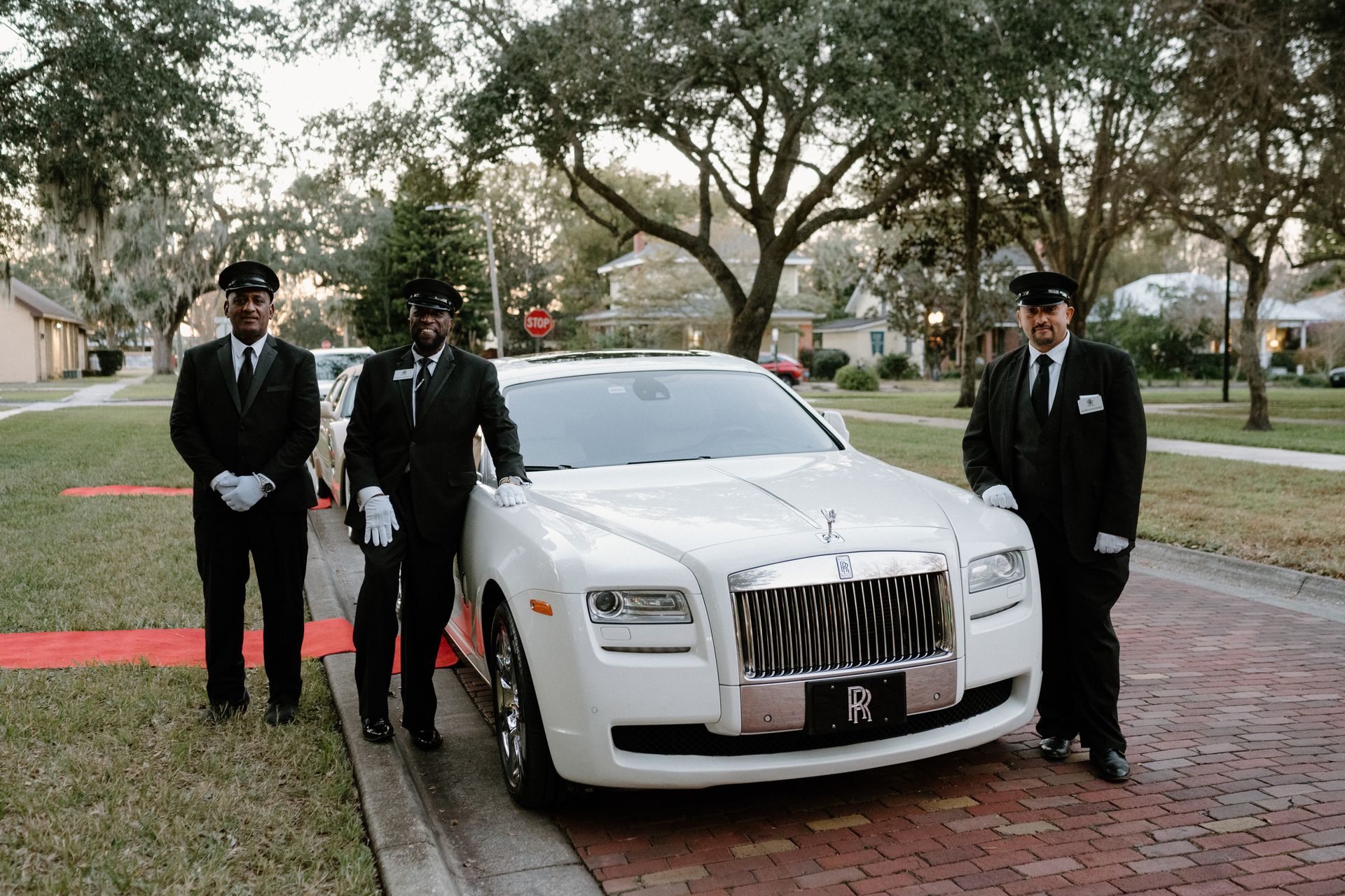 Three chauffeurs in formal black suits and white gloves stand beside a white Rolls-Royce parked on a brick street.