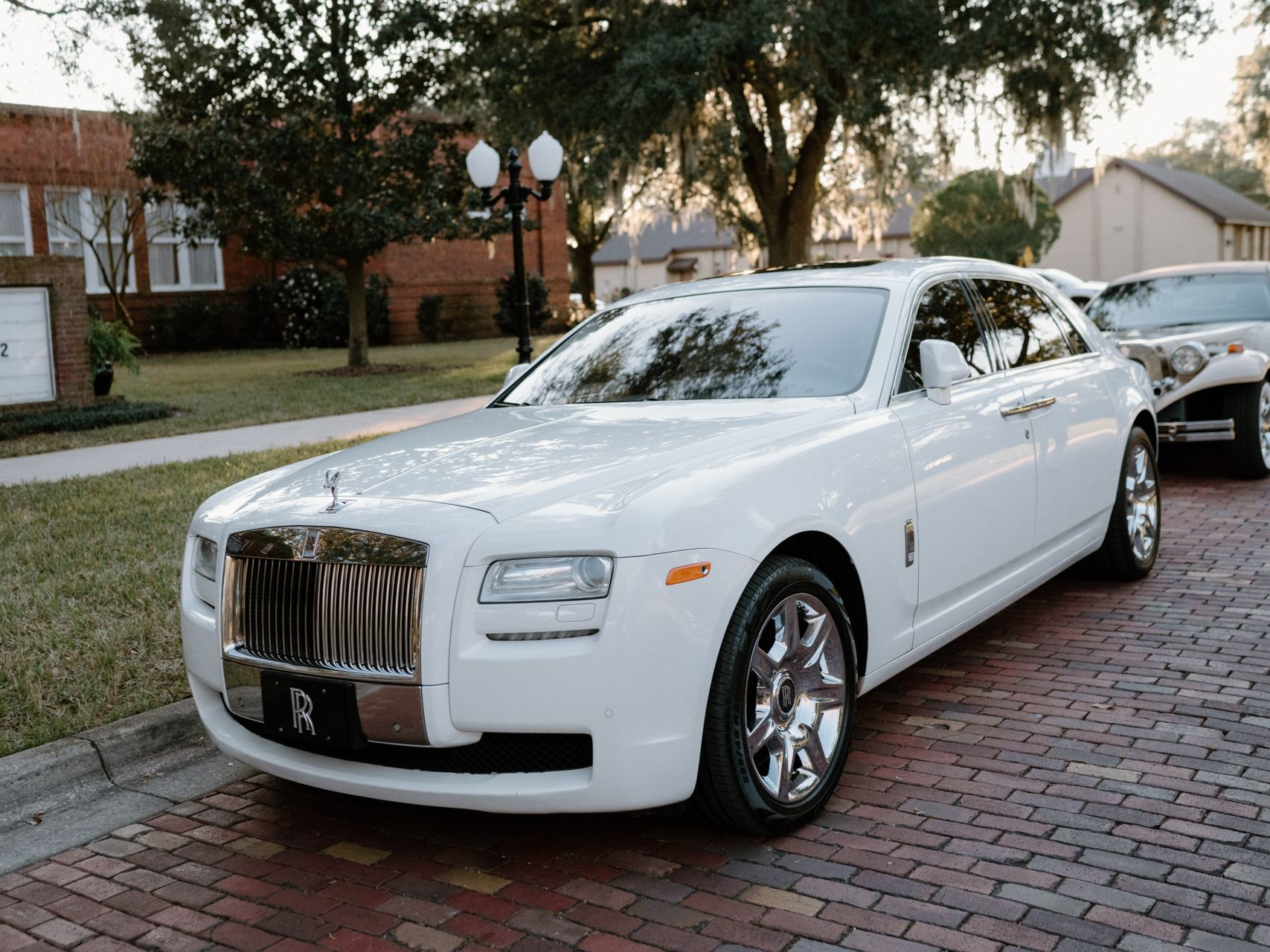 A white Rolls-Royce Ghost parked on a brick road near a lawn and building.