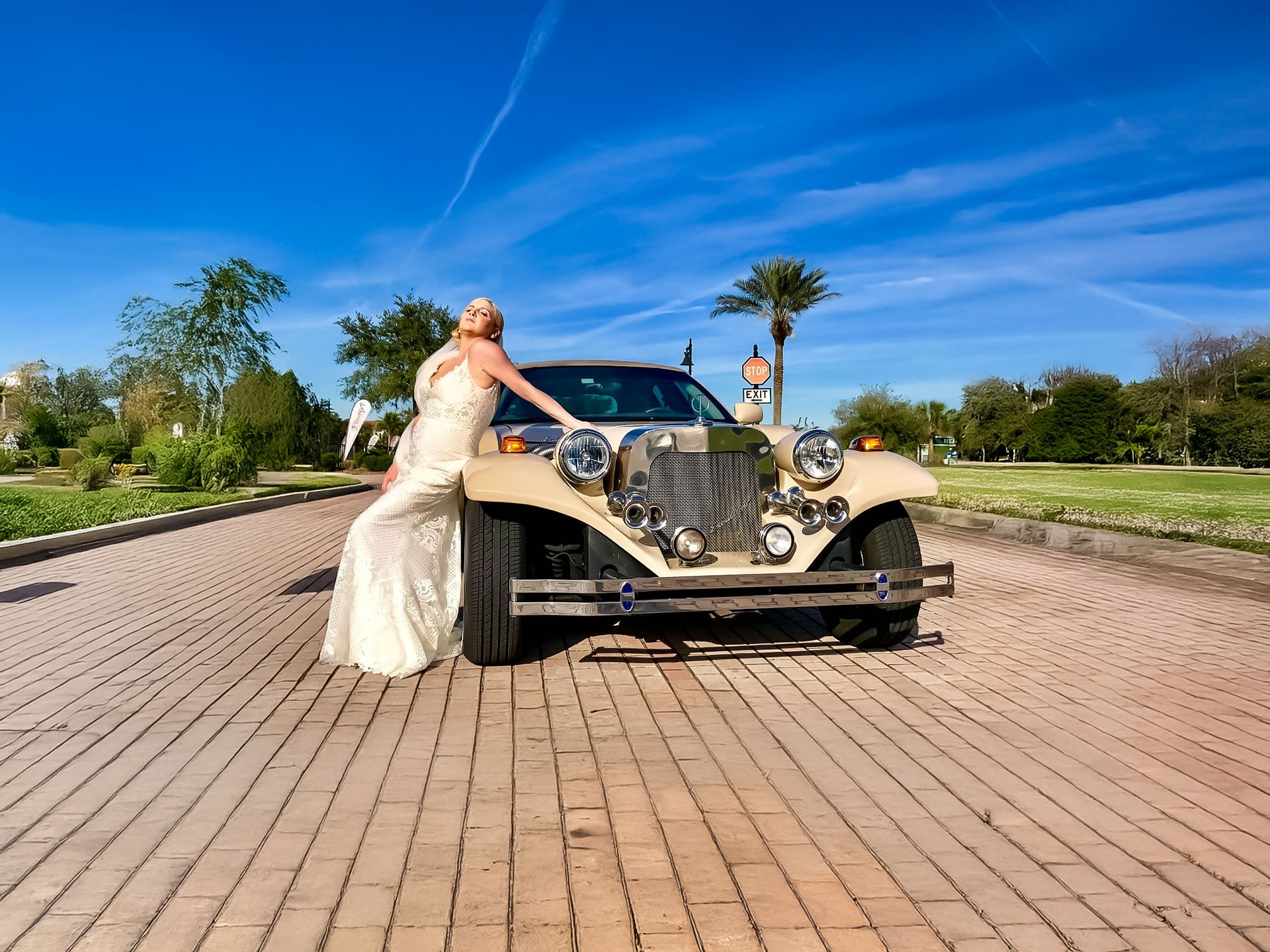 A person in a long, light-colored dress poses against a beige vintage-style car on a brick path under a sunny blue sky.