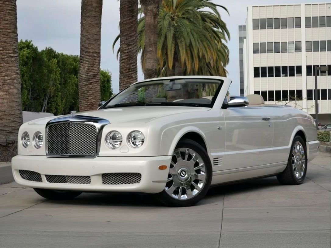 A white Bentley convertible parked on a sidewalk with palm trees and a modern office building in the background.