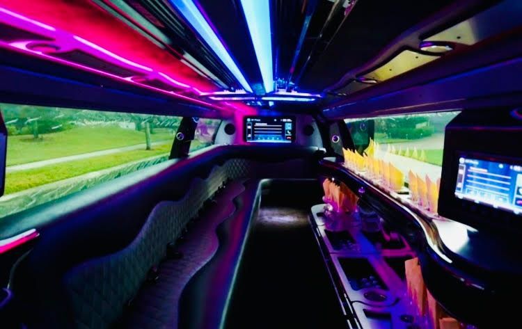 The interior of a luxury limousine featuring plush seating, vibrant purple and blue neon lighting, and digital screens.