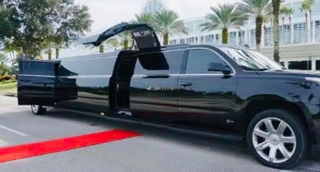 A black stretch limousine with gull-wing doors open, parked outdoors on a paved lot next to a red carpet.