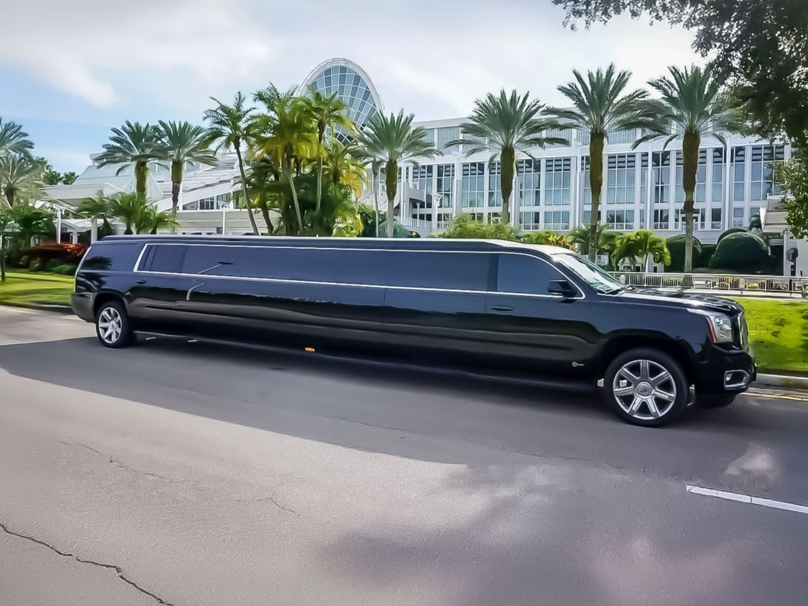 A long, black stretch limousine parked on a road in front of a white building with palm trees.
