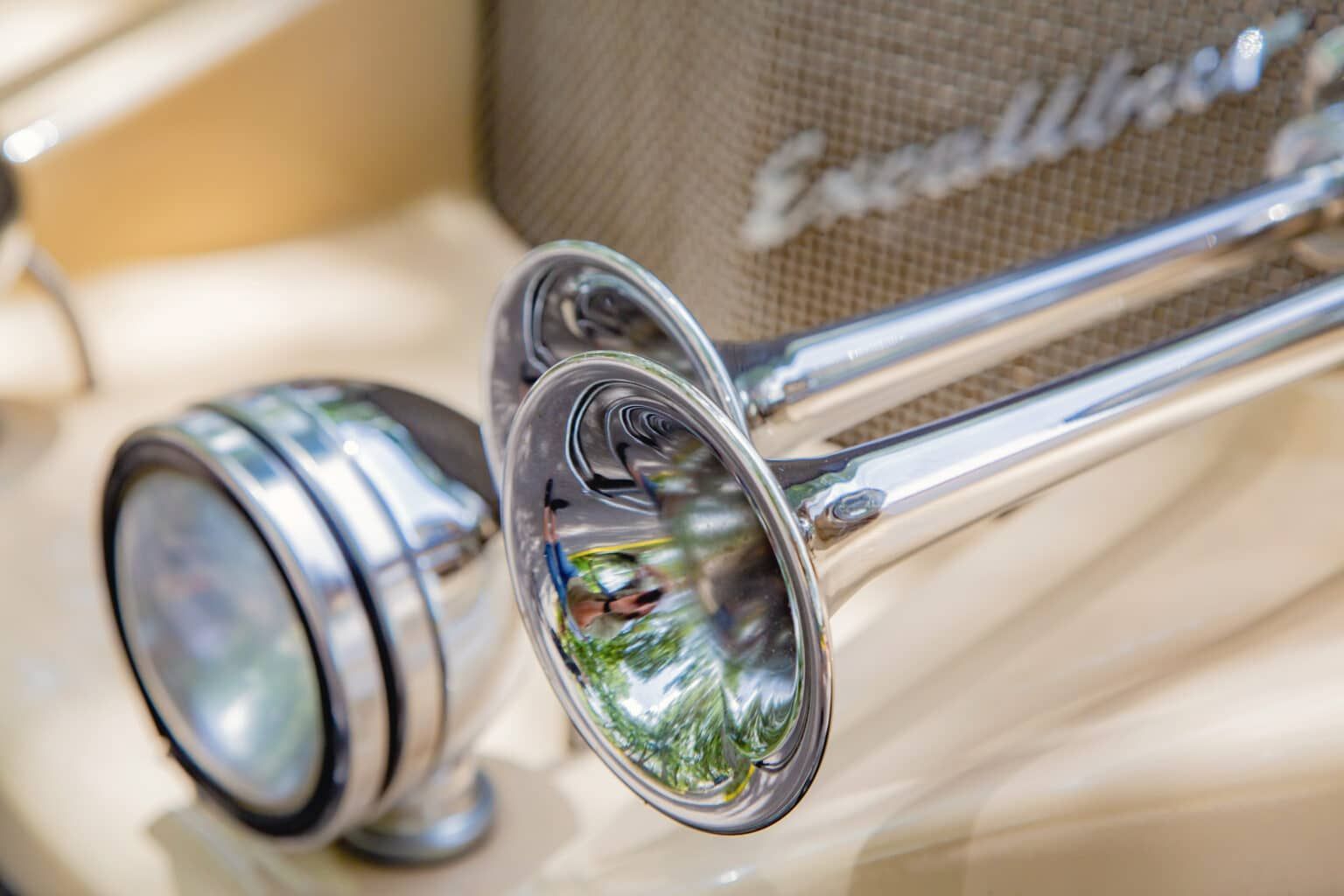 Chrome trumpet horns and a round headlight on the tan, glossy body of a vintage Excalibur car.