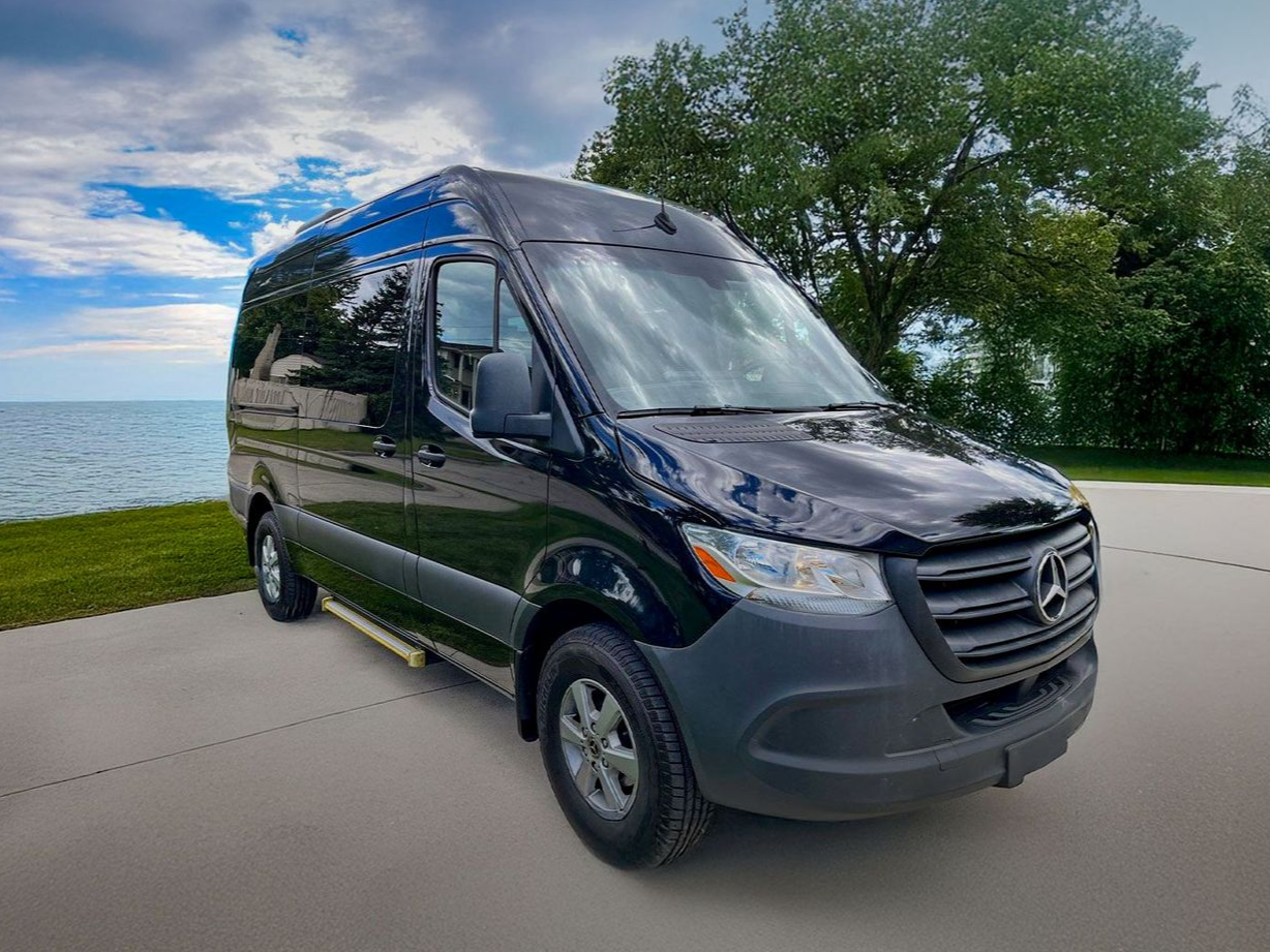 A black Mercedes-Benz Sprinter van parked on a paved lot beside a body of water with trees in the background.