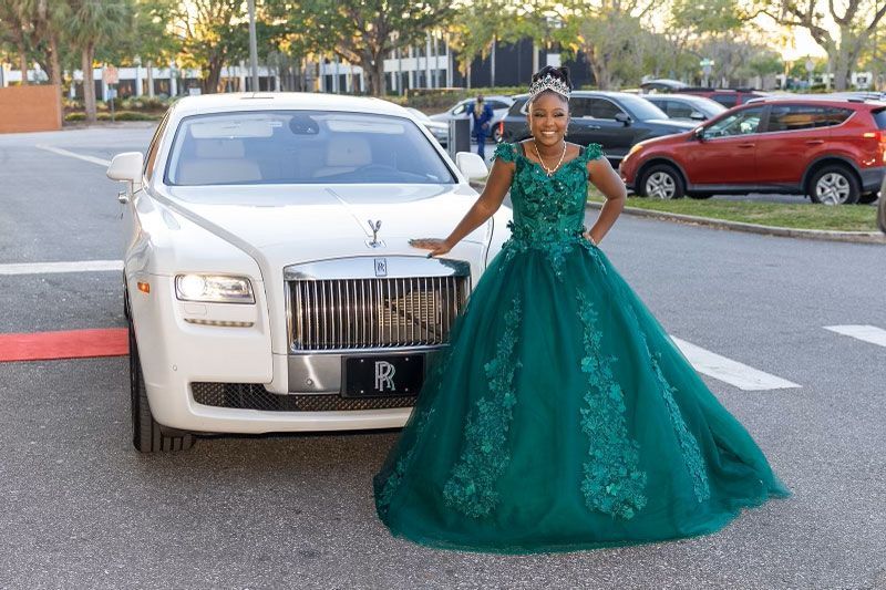 A person in a dark green, floor-length ballgown stands smiling next to a white Rolls-Royce parked in a lot.