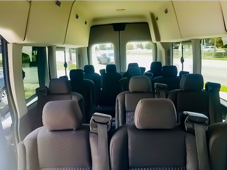 Interior view of a passenger van with multiple rows of dark gray fabric seats facing the rear doors.