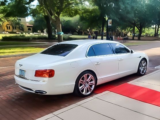 A white Bentley sedan parked on a brick path next to a red carpet, with trees and a building in the background.