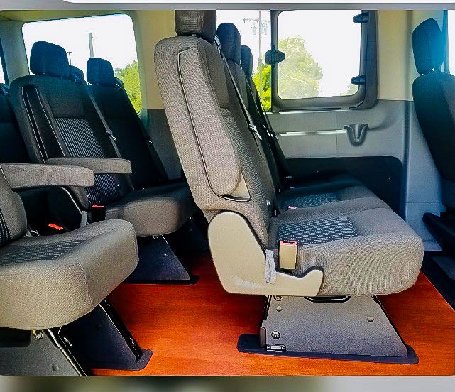 Interior of a passenger van featuring rows of grey upholstered seats with armrests on a polished wood-look floor.