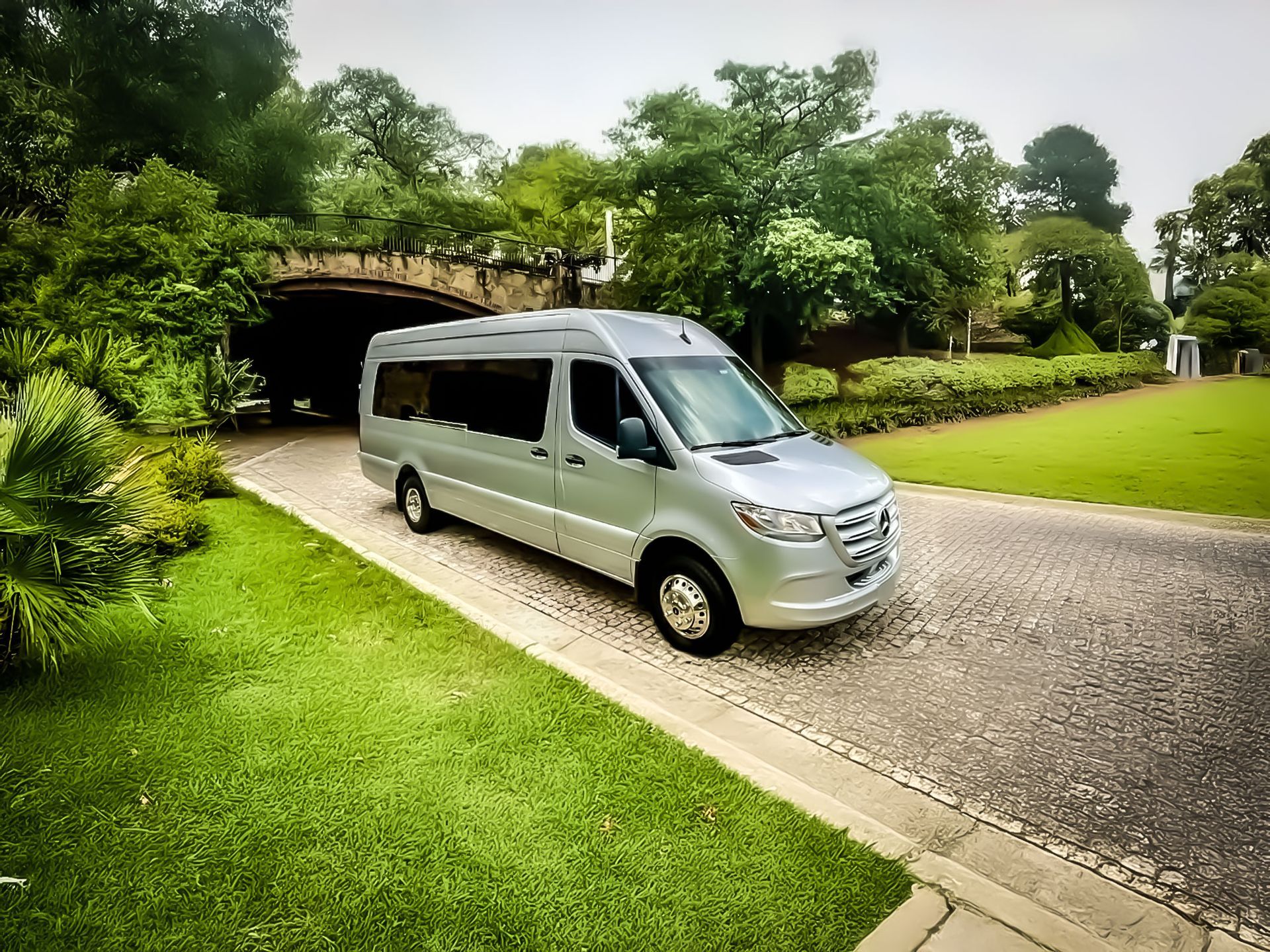 A silver passenger van driving on a paved road through a park with a stone bridge and green lawns in the background.