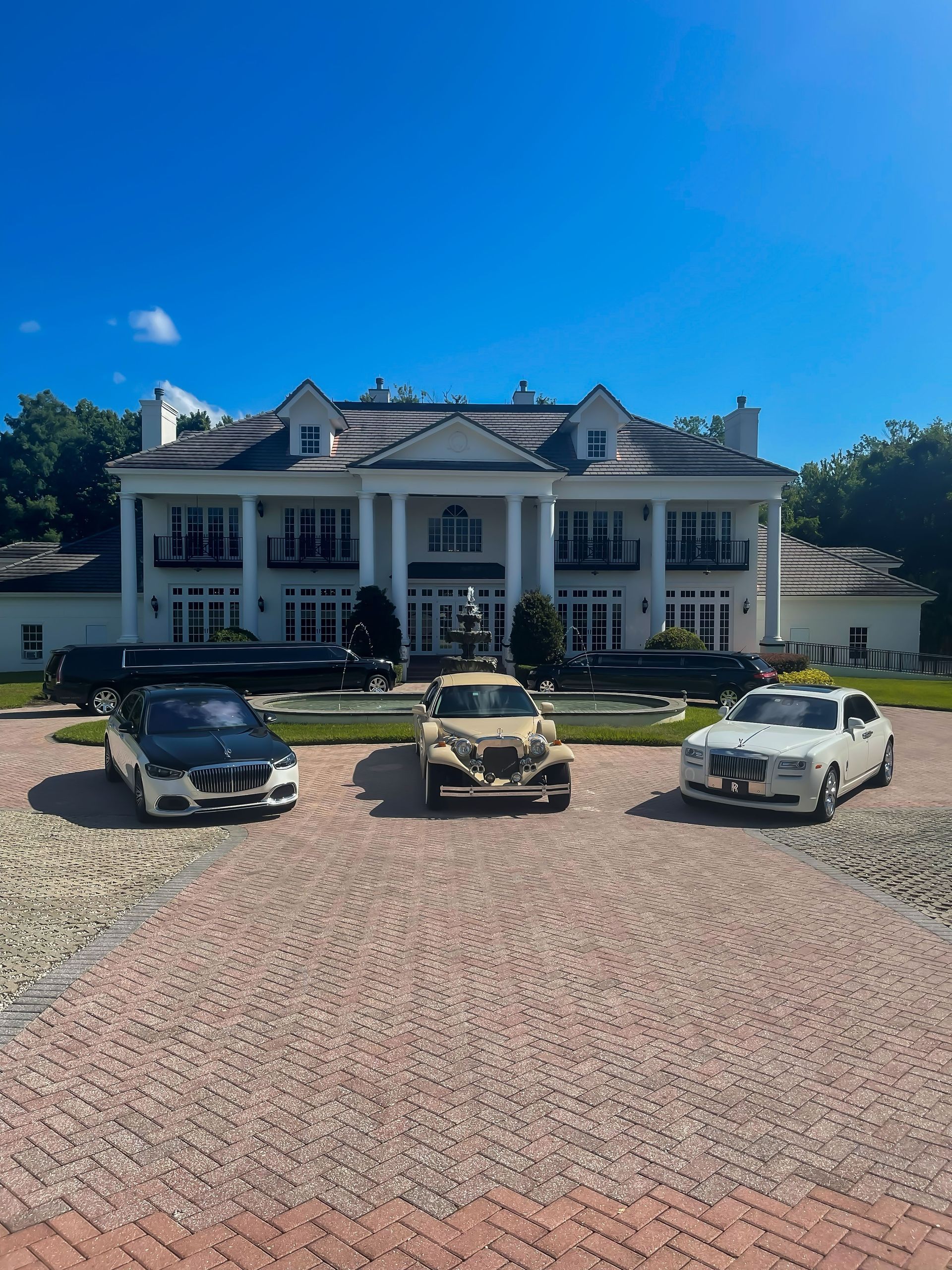 Three luxury cars parked in a driveway in front of a grand, white, two-story colonial-style mansion under a clear blue sky.