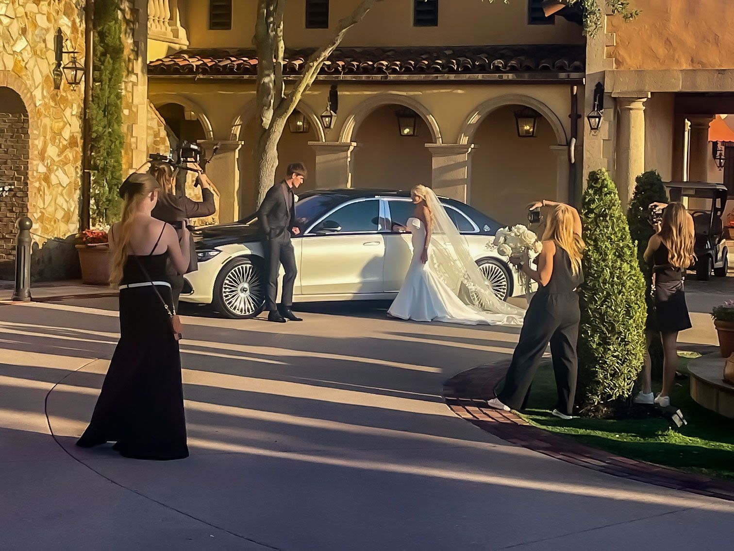 A wedding photoshoot with a bride in a white gown and a man by a car, with two photographers on a stone plaza.