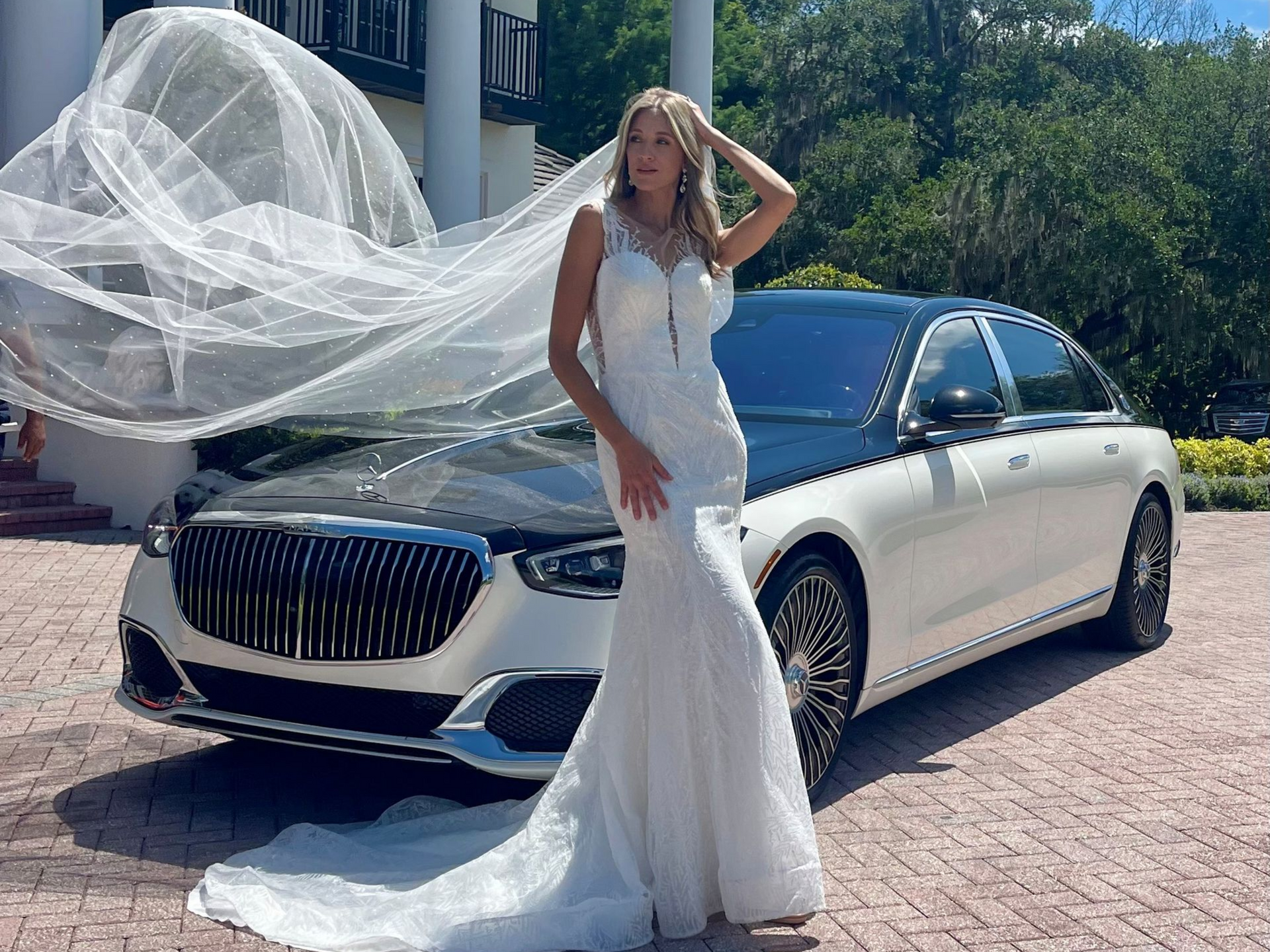 A person in a white wedding gown and long flowing veil poses in front of a two-tone luxury sedan on a paved driveway.
