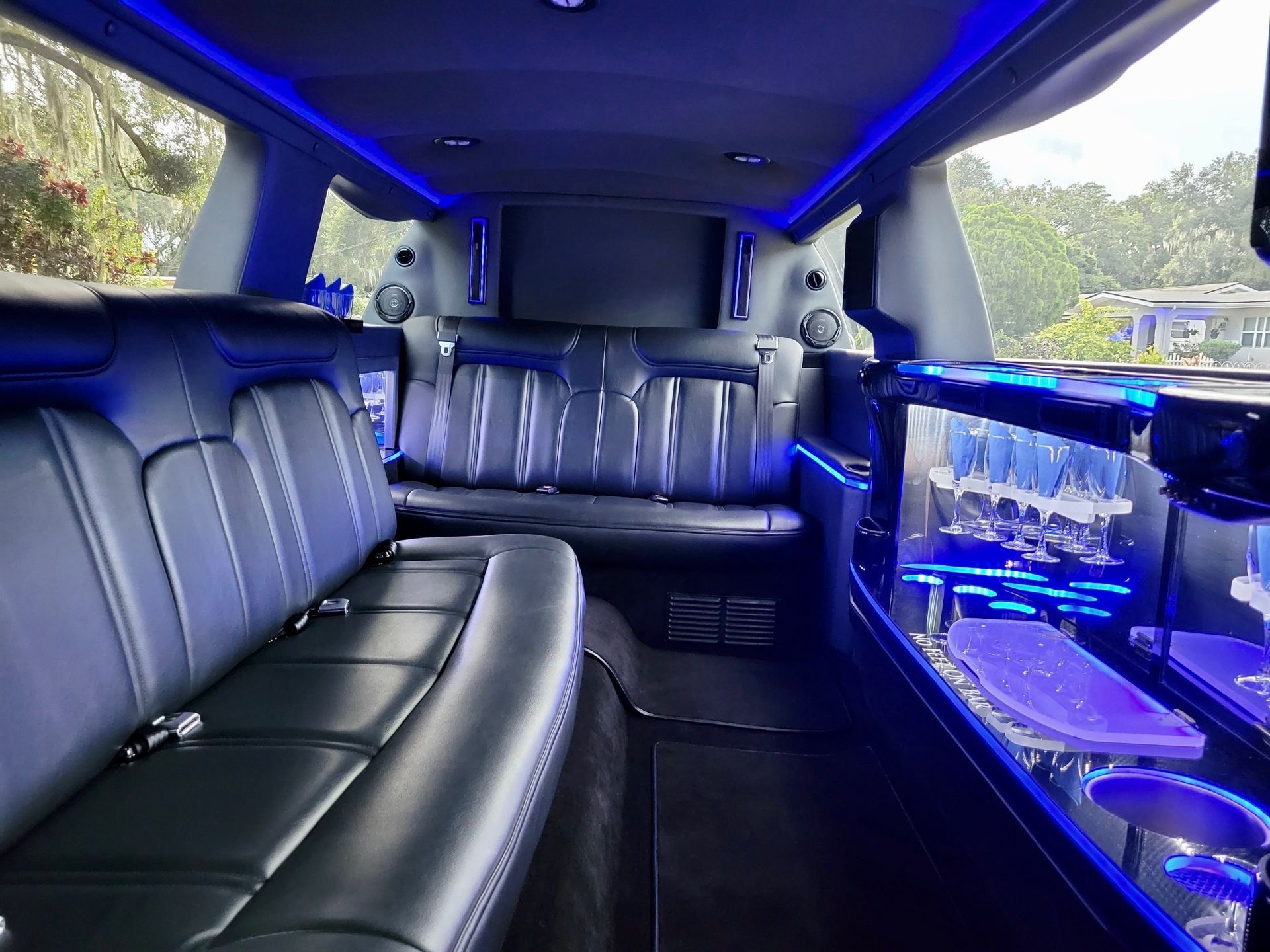 The interior of a limousine featuring black leather bench seating, blue ambient lighting, and a glass-filled bar area.