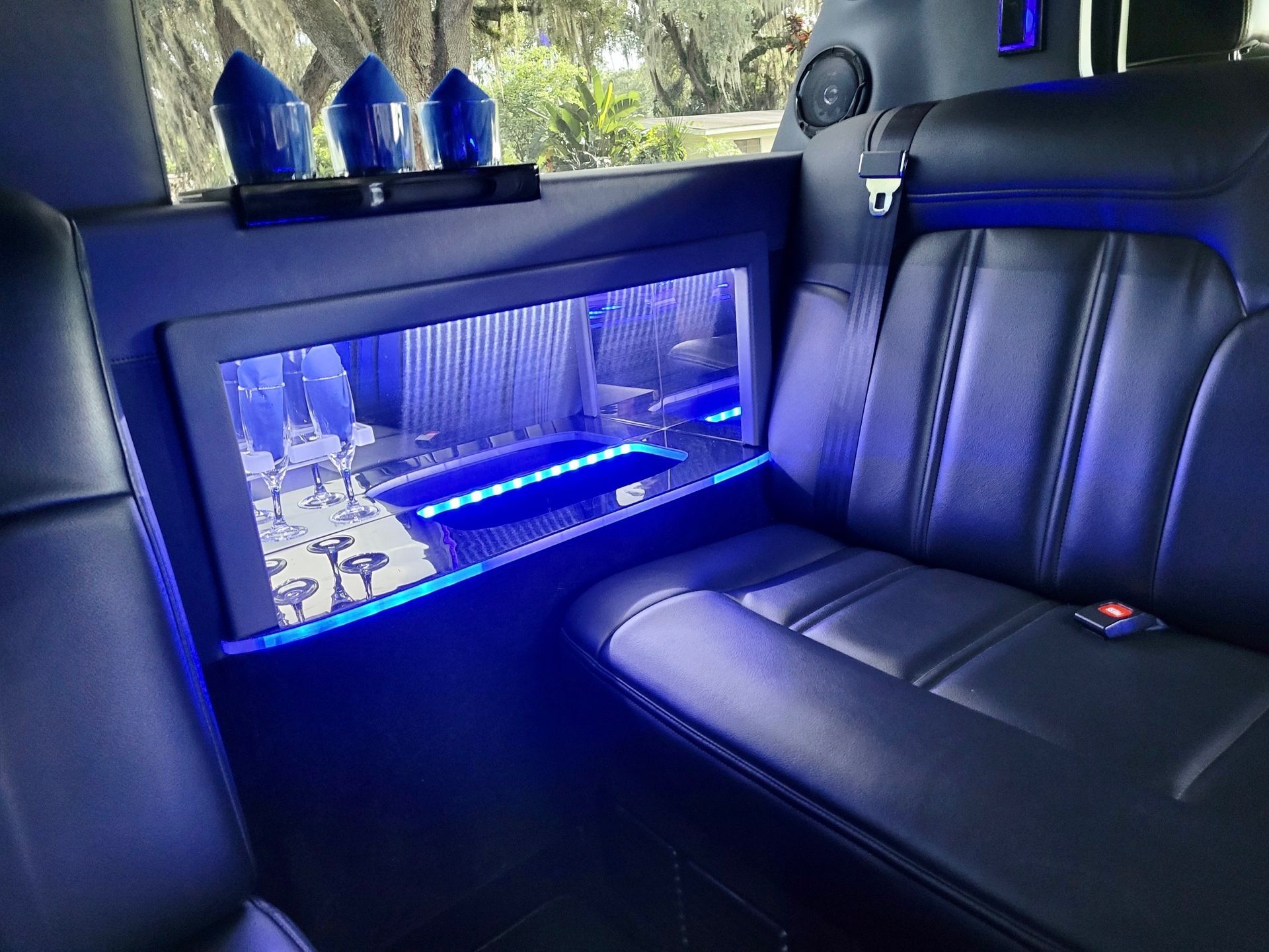 Interior of a limousine with dark leather seats, blue ambient lighting, and a drink shelf with three blue glasses.