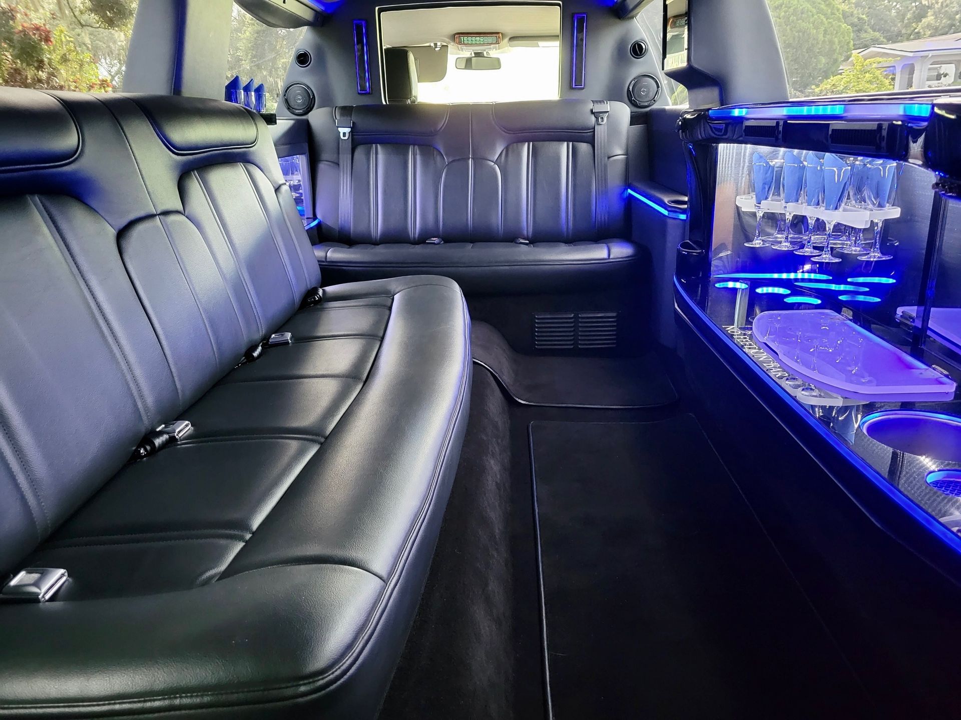 The interior of a black luxury limousine with leather bench seating, purple accent lighting, and a drink bar.