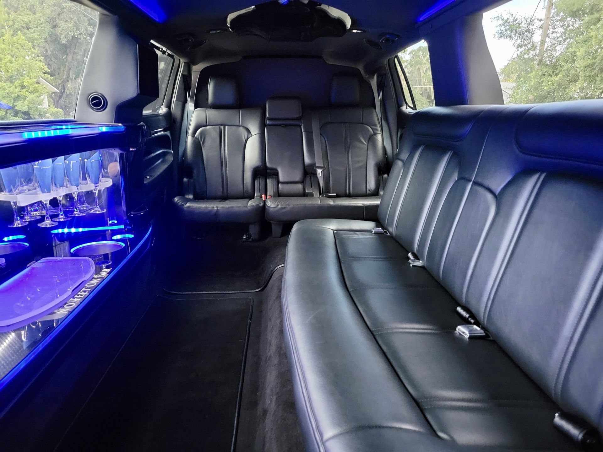 Interior of a luxury limousine with black leather seats, blue accent lighting, and a built-in bar area.